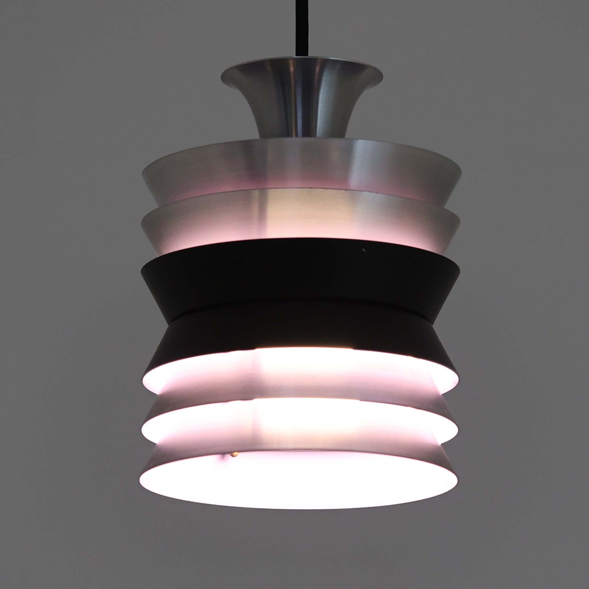 Carl ThoreがGranhaga Metallindustriで手掛けた直径15cmの小ぶりなペンダントライトで、段状に重ねたアルミシェードが光を柔らかく拡散し、点灯時には上下方向へ美しいグラデーションを生み出す北欧ビンテージ品です。繊細なヘアライン仕上げとブラックパーツのコントラストがモダンな印象を与え、キッチンカウンターや小空間のアクセント照明としてはもちろん、複数灯を並べたコーディネートにも最適です。コンパクトながら彫刻的なフォルムが存在感を放ち、経年で深まった質感がビンテージならではの魅力を際立たせています。