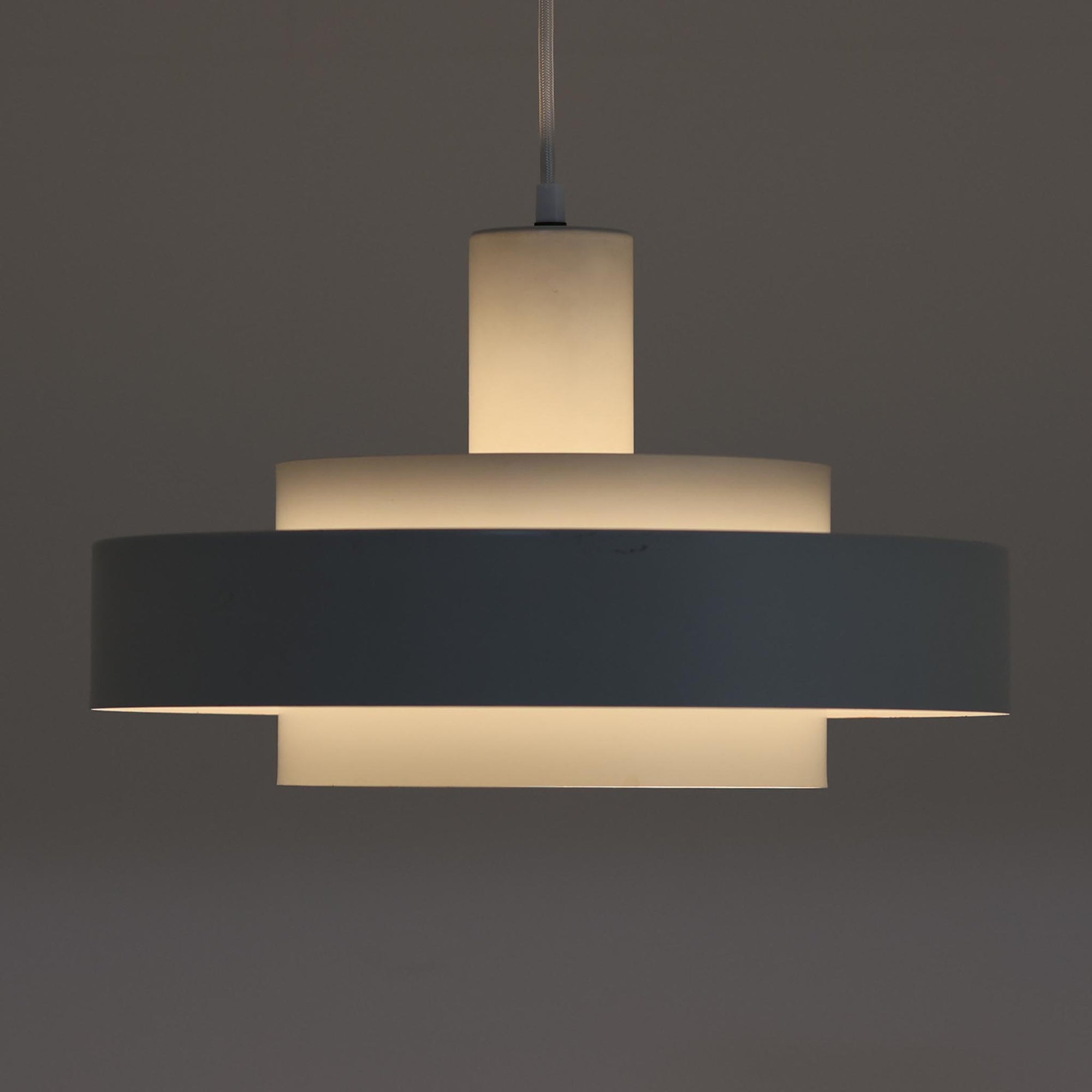 Jo HammerborgがFog & Morupのためにデザインした「Equator」は、直径40cmの存在感あるスチール製ペンダントライトで、赤・黄色・青など多彩なカラーバリエーションが展開されていたモデルとして知られますが、本品は空間に優しく溶け込む上品なホワイトカラーが採用されています。スチール特有の程よい重量感と重厚な佇まいがあり、実際に吊るすとホワイトの柔らかな印象の中にしっかりとした質感が感じられるのも魅力です。同心円状に重なるシェードが光を柔らかく分散させ、眩しさを抑えつつ立体的な陰影を生み出す構造が特徴で、ダイニングやリビングの主照明として落ち着きのあるモダンなアクセントを添えます。ミニマルな造形の中に職人技が凝縮されており、経年で深まった塗装の質感がビンテージならではの魅力を一層引き立てています。