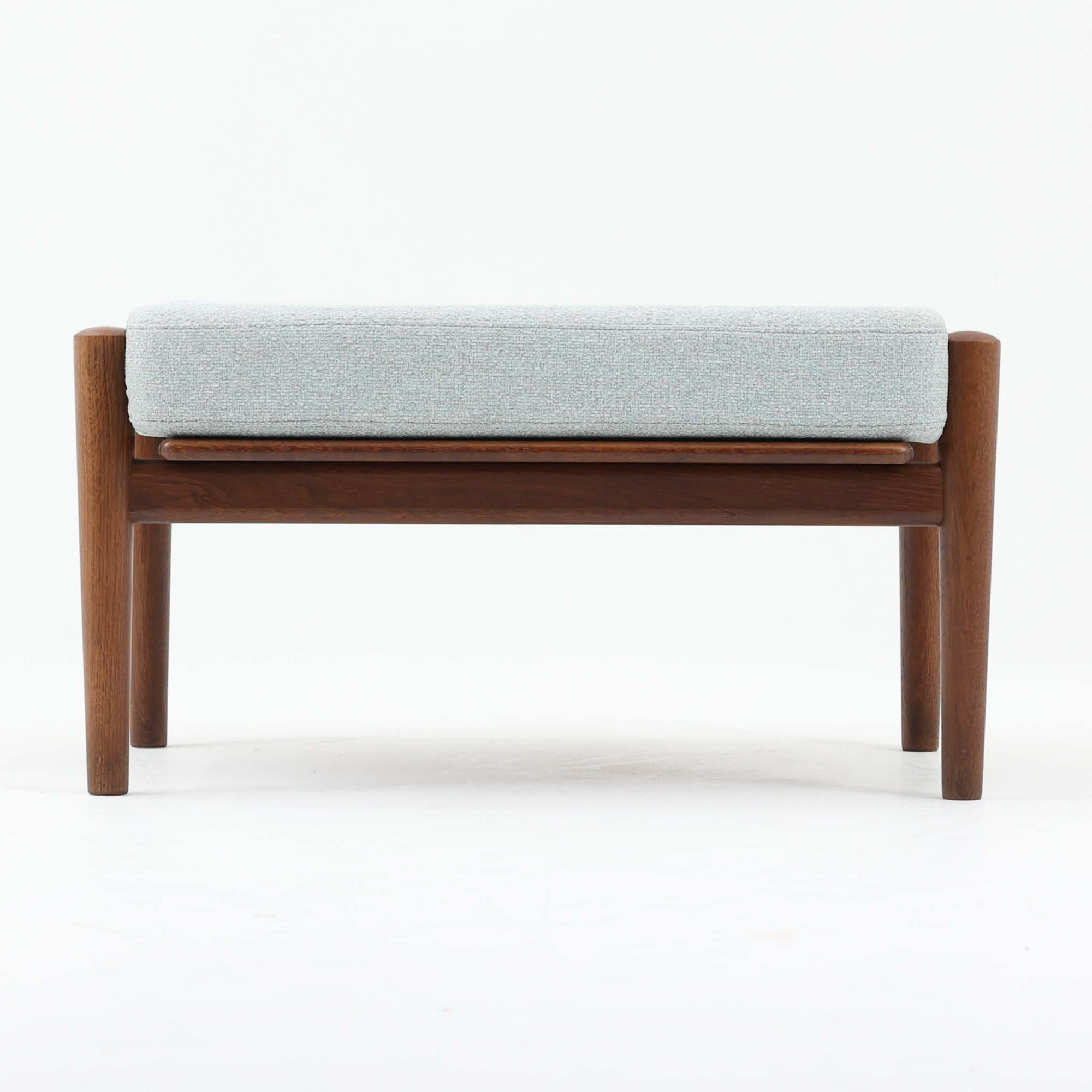 Hans J. Wegnerがデザインし、Getamaが製作したGE530オットマンです。ダークオーク材の力強い木目を活かした角の取れたフレームと、丸脚を組み合わせた端正な構造が特徴で、裏面には刻印も確認できます。程よい厚みのクッションは張り替え済みで、軽やかな生地が木部の深みのある色合いを引き立てます。同シリーズのラウンジチェアと合わせてはもちろん、単体でスツールや補助椅子としても活躍する、北欧ビンテージならではの質実剛健な一品です。