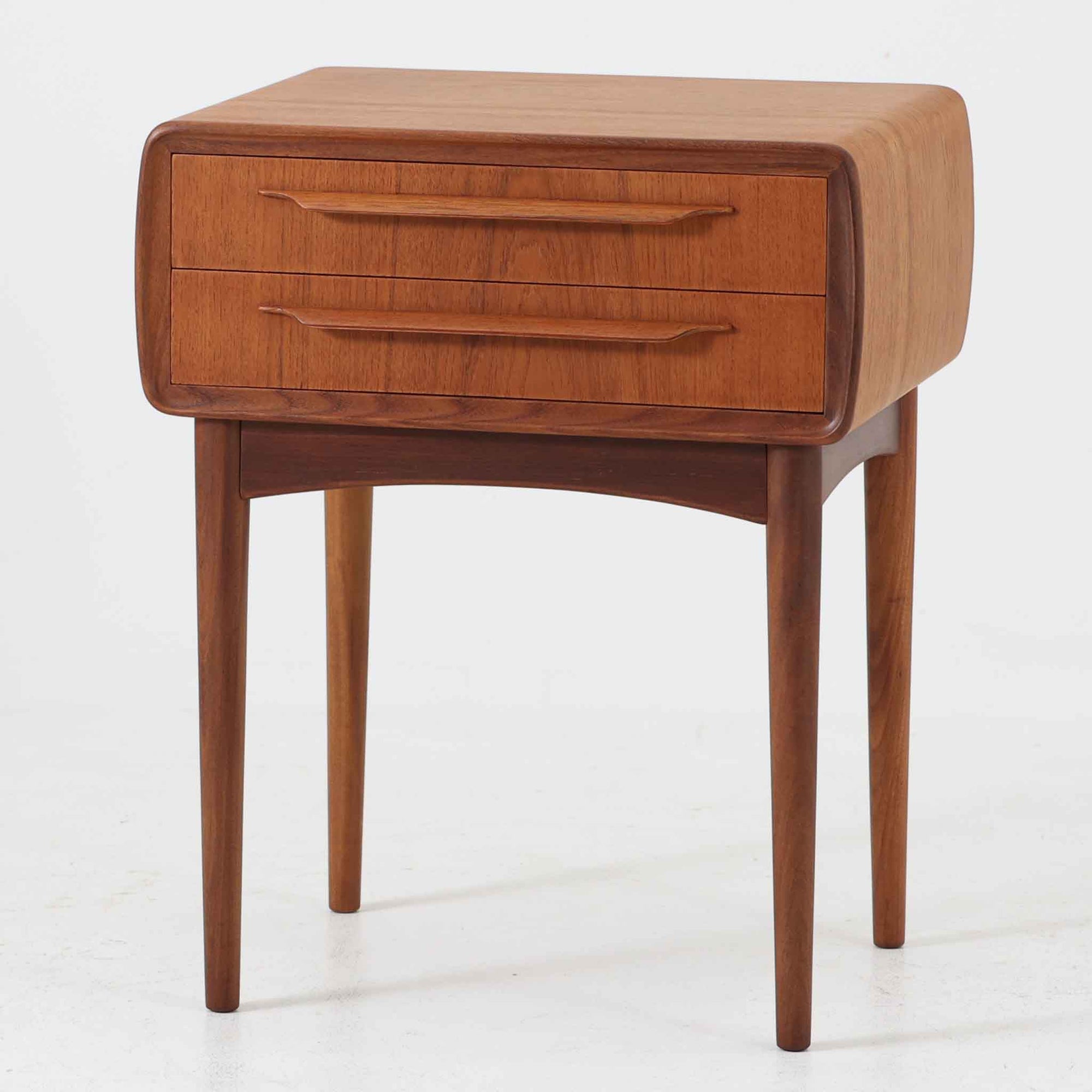 Johannes Andersen（ヨハネス・アンダーセン）がCFC Silkeborgのために手掛けた2段チェストは、チーク材を贅沢に用いた柔らかな曲面フォルムと長く伸びた彫刻的なハンドルが印象的で、天板から側面へと美しくつながる木目が上質な素材感を際立たせる北欧ビンテージ家具です。職人技の光る滑らかな面取りや端正な造りが深い味わいを生み出し、コンパクトながら実用的な2杯の引き出しを備え、ベッドサイドやリビングのサイドテーブルとしても活躍し、温かみのある木肌の経年変化が魅力を一層引き立てています。