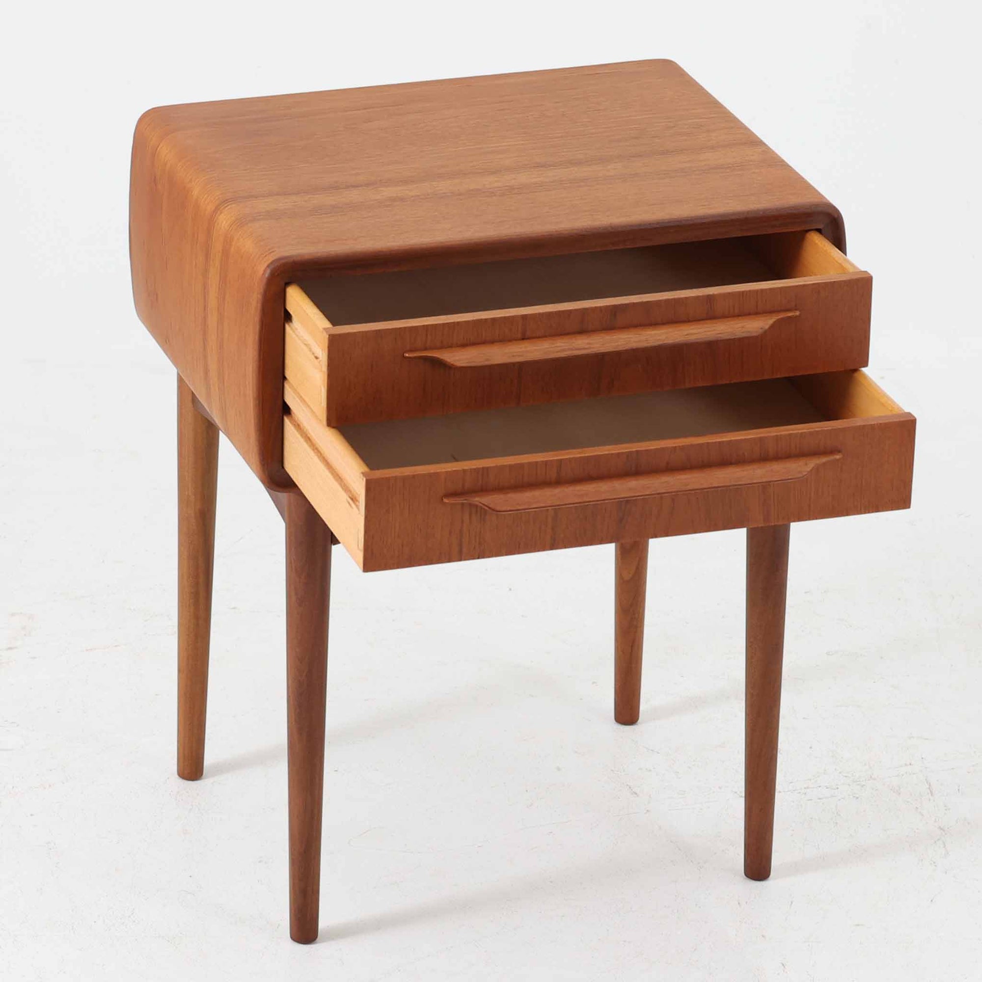 Johannes Andersen（ヨハネス・アンダーセン）がCFC Silkeborgのために手掛けた2段チェストは、チーク材を贅沢に用いた柔らかな曲面フォルムと長く伸びた彫刻的なハンドルが印象的で、天板から側面へと美しくつながる木目が上質な素材感を際立たせる北欧ビンテージ家具です。職人技の光る滑らかな面取りや端正な造りが深い味わいを生み出し、コンパクトながら実用的な2杯の引き出しを備え、ベッドサイドやリビングのサイドテーブルとしても活躍し、温かみのある木肌の経年変化が魅力を一層引き立てています。