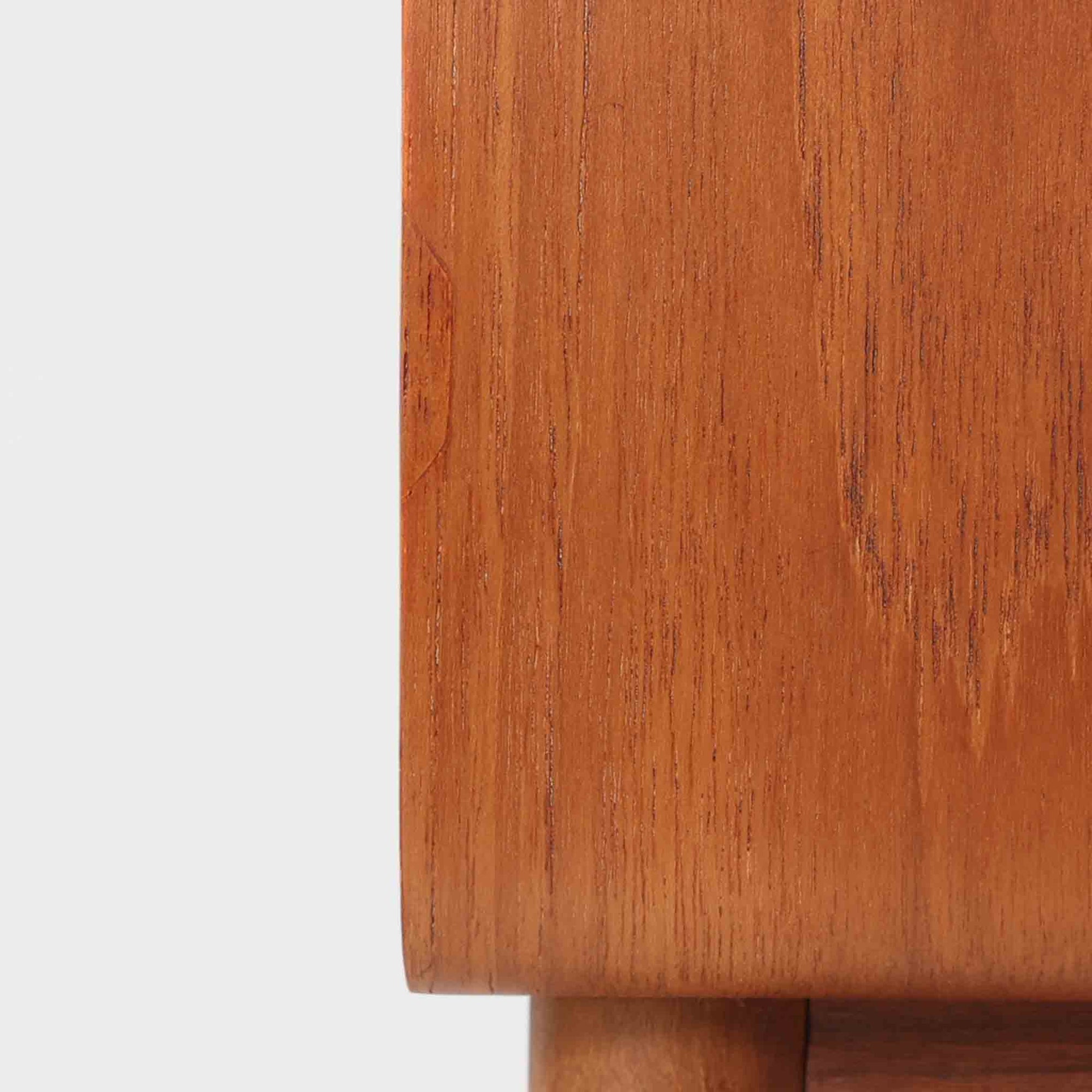 Johannes Andersen（ヨハネス・アンダーセン）がCFC Silkeborgのために手掛けた2段チェストは、チーク材を贅沢に用いた柔らかな曲面フォルムと長く伸びた彫刻的なハンドルが印象的で、天板から側面へと美しくつながる木目が上質な素材感を際立たせる北欧ビンテージ家具です。職人技の光る滑らかな面取りや端正な造りが深い味わいを生み出し、コンパクトながら実用的な2杯の引き出しを備え、ベッドサイドやリビングのサイドテーブルとしても活躍し、温かみのある木肌の経年変化が魅力を一層引き立てています。