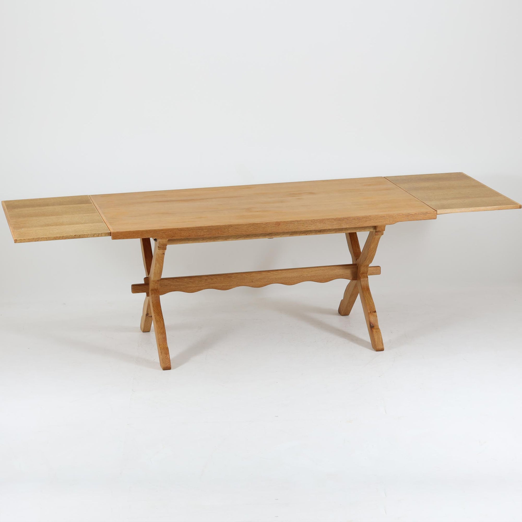 Henning Kjaernulfがデザインし、Nyrup Mobelfabrikが手掛けたエクステンションダイニングテーブルです。オーク材ならではの力強い木目と経年で深みを増した色艶が魅力で、X字状の脚部や波状に削り出された貫が北欧らしい素朴さと彫刻的な美しさを演出しています。左右の天板を引き出すことで幅251cmまで拡張可能な実用的構造で、日常使いから来客時まで幅広く活躍。重厚感がありながらも軽やかな意匠にまとめられた、希少性の高い北欧ビンテージ家具です。
