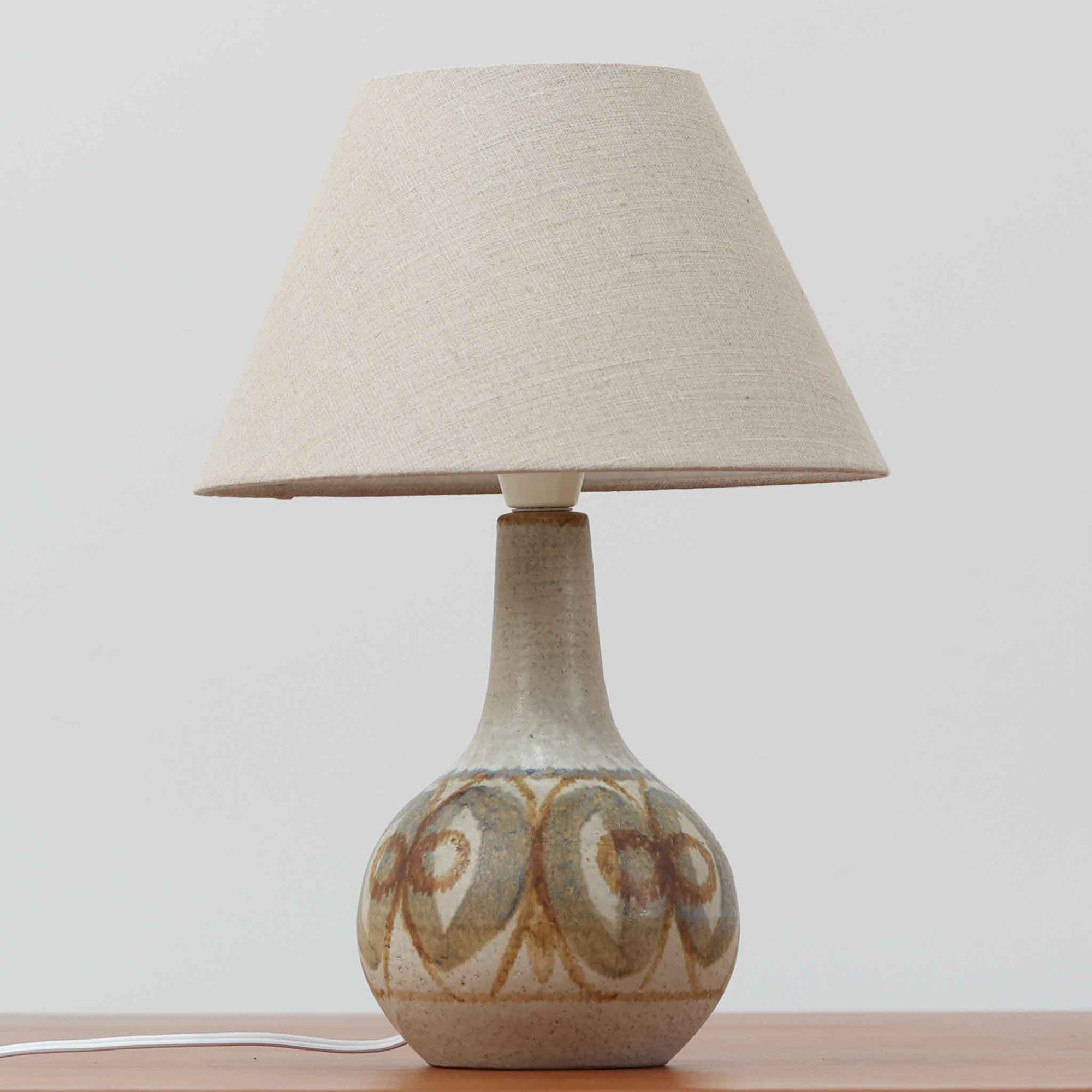 スーホルム・セラミック（Soholm Keramik）によるこちらのテーブルランプは、温かみのある陶磁器ベースに柔らかなブラウンとブルーで描かれた連続模様が印象的で、どこか手仕事の息づかいを感じさせる北欧ビンテージらしい魅力を備えています。丸みを帯びたベースからすっと伸びる首部のラインが美しく、リネンシェードを通した穏やかな灯りが空間にやさしい陰影を生み出します。高さ35cmと日常使いしやすいサイズで、ベッドサイドやリビングのサイドテーブルにも程よく収まり、落ち着いたインテリアに自然に馴染む一台です。