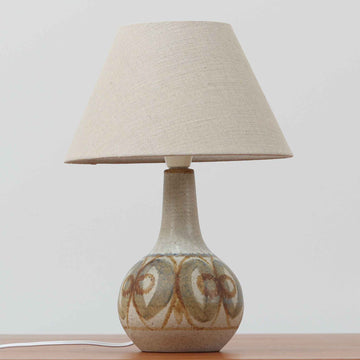 スーホルム・セラミック（Soholm Keramik）によるこちらのテーブルランプは、温かみのある陶磁器ベースに柔らかなブラウンとブルーで描かれた連続模様が印象的で、どこか手仕事の息づかいを感じさせる北欧ビンテージらしい魅力を備えています。丸みを帯びたベースからすっと伸びる首部のラインが美しく、リネンシェードを通した穏やかな灯りが空間にやさしい陰影を生み出します。高さ35cmと日常使いしやすいサイズで、ベッドサイドやリビングのサイドテーブルにも程よく収まり、落ち着いたインテリアに自然に馴染む一台です。