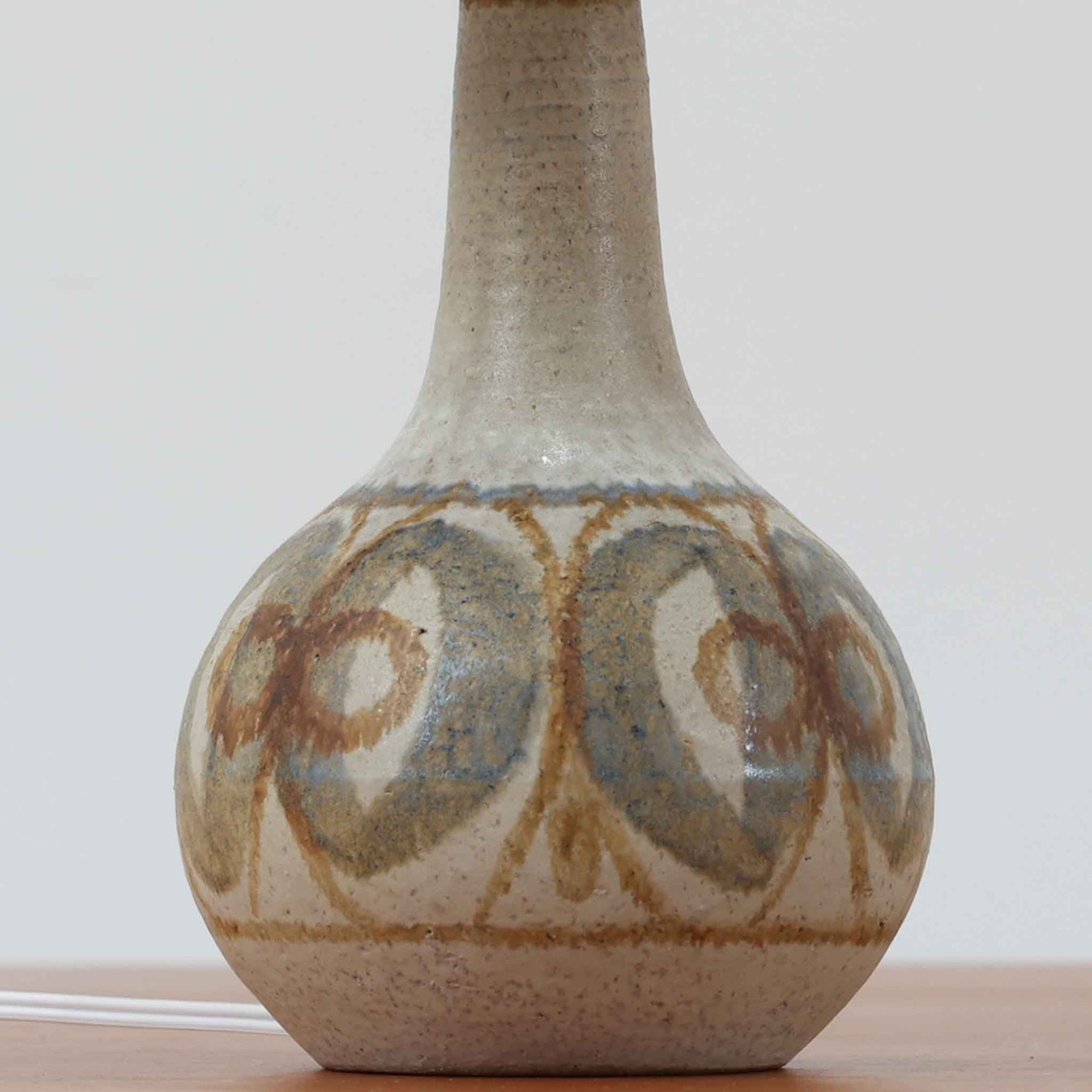 スーホルム・セラミック（Soholm Keramik）によるこちらのテーブルランプは、温かみのある陶磁器ベースに柔らかなブラウンとブルーで描かれた連続模様が印象的で、どこか手仕事の息づかいを感じさせる北欧ビンテージらしい魅力を備えています。丸みを帯びたベースからすっと伸びる首部のラインが美しく、リネンシェードを通した穏やかな灯りが空間にやさしい陰影を生み出します。高さ35cmと日常使いしやすいサイズで、ベッドサイドやリビングのサイドテーブルにも程よく収まり、落ち着いたインテリアに自然に馴染む一台です。