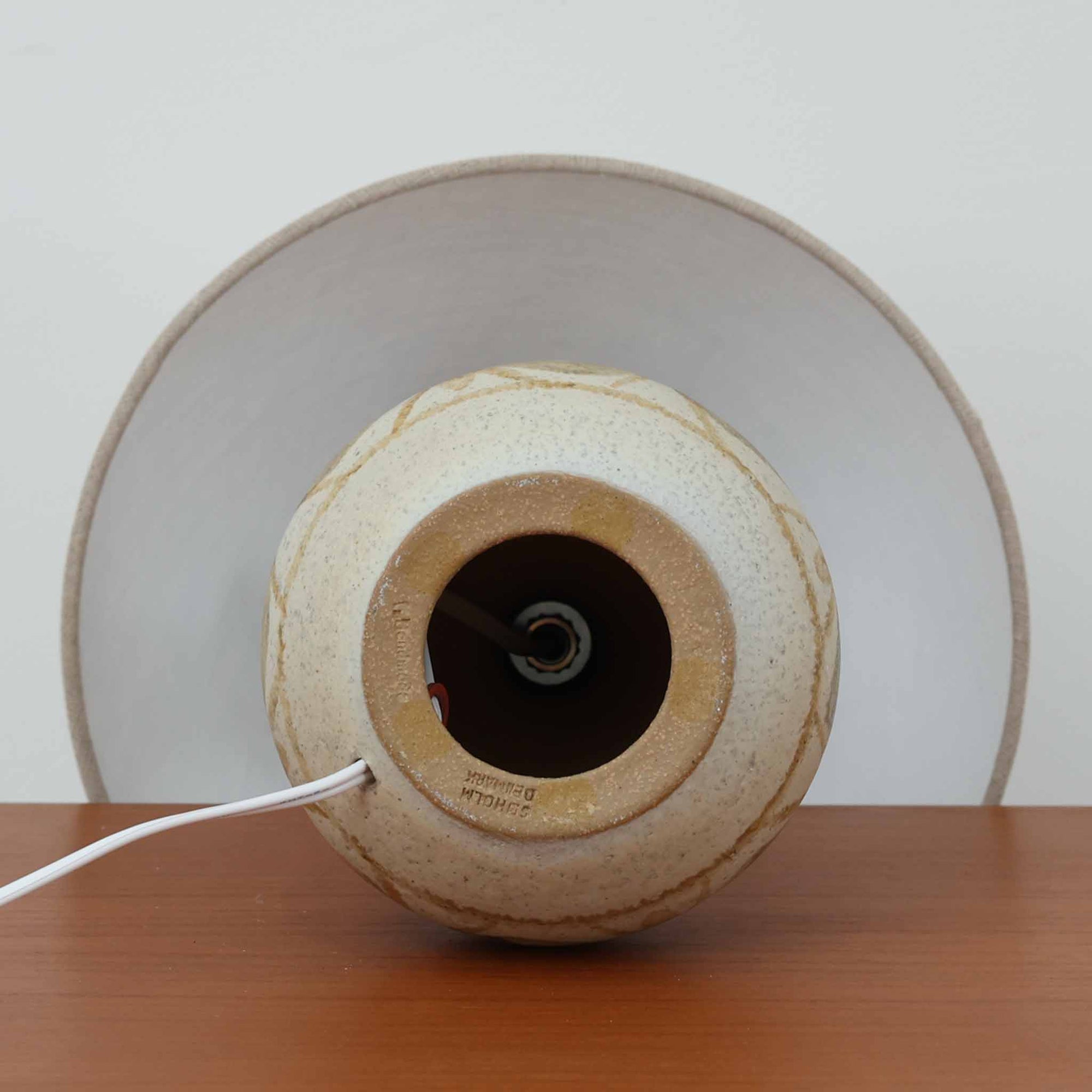 スーホルム・セラミック（Soholm Keramik）によるこちらのテーブルランプは、温かみのある陶磁器ベースに柔らかなブラウンとブルーで描かれた連続模様が印象的で、どこか手仕事の息づかいを感じさせる北欧ビンテージらしい魅力を備えています。丸みを帯びたベースからすっと伸びる首部のラインが美しく、リネンシェードを通した穏やかな灯りが空間にやさしい陰影を生み出します。高さ35cmと日常使いしやすいサイズで、ベッドサイドやリビングのサイドテーブルにも程よく収まり、落ち着いたインテリアに自然に馴染む一台です。