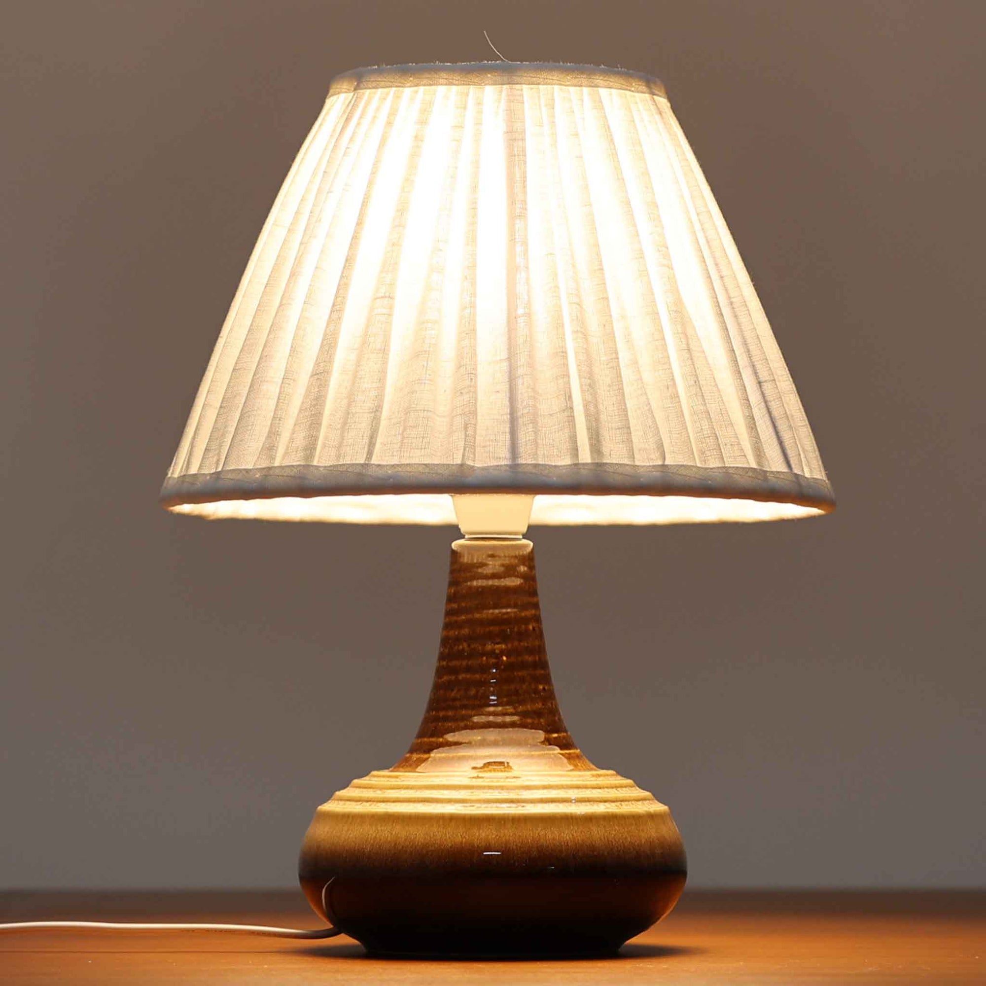 Soholm Keramik（スーホルム・セラミック）が手掛けたモデル1214のテーブルランプは、艶やかなブラウンの釉薬と段状のレリーフが生む豊かな表情が魅力で、陶磁器ならではの重量感と温かみが空間に落ち着いた存在感をもたらす北欧ビンテージ品です。柔らかな光を透過するリネンシェードがやさしい灯りを広げ、ベッドサイドやリビングのアクセント照明として取り入れやすく、経年の風合いが一層味わいを深めています。
