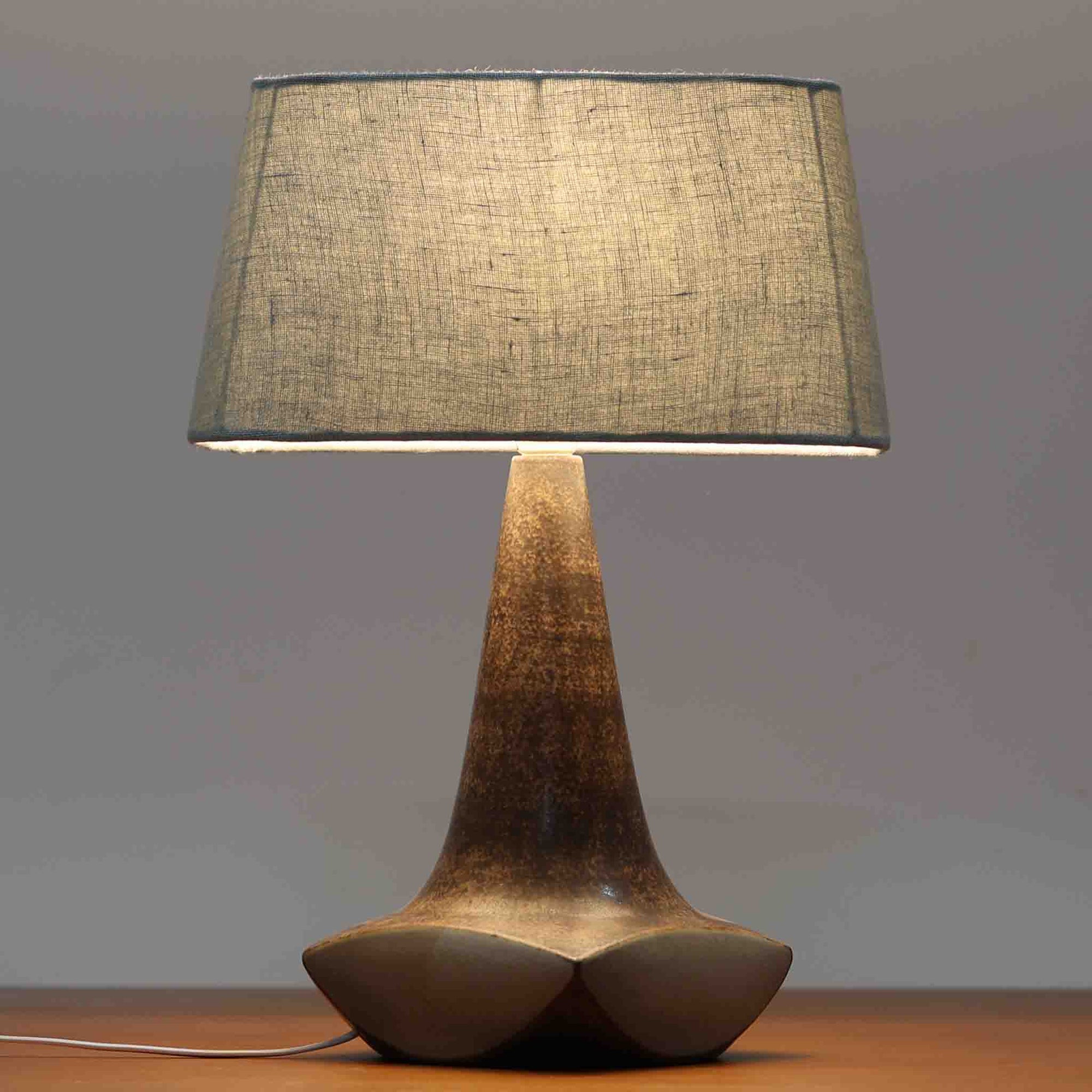 Michael Andersen & Sonが手掛けたこちらのテーブルランプは、陶磁器ならではの落ち着いた質感と、彫刻的に立ち上がるフォルムが存在感を放つ北欧ビンテージ品で、マットブラウンから生成り色へと移ろう釉薬のグラデーションが温かみある陰影を生み出し、リネンシェードを通したやわらかな灯りがベッドサイドやリビングに静かな安らぎを添えます。経年で深まった陶肌の表情がひとつとして同じものがない魅力となり、空間のアクセント照明として長くお楽しみいただける一台です。