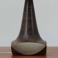 Michael Andersen & Sonが手掛けたこちらのテーブルランプは、陶磁器ならではの落ち着いた質感と、彫刻的に立ち上がるフォルムが存在感を放つ北欧ビンテージ品で、マットブラウンから生成り色へと移ろう釉薬のグラデーションが温かみある陰影を生み出し、リネンシェードを通したやわらかな灯りがベッドサイドやリビングに静かな安らぎを添えます。経年で深まった陶肌の表情がひとつとして同じものがない魅力となり、空間のアクセント照明として長くお楽しみいただける一台です。