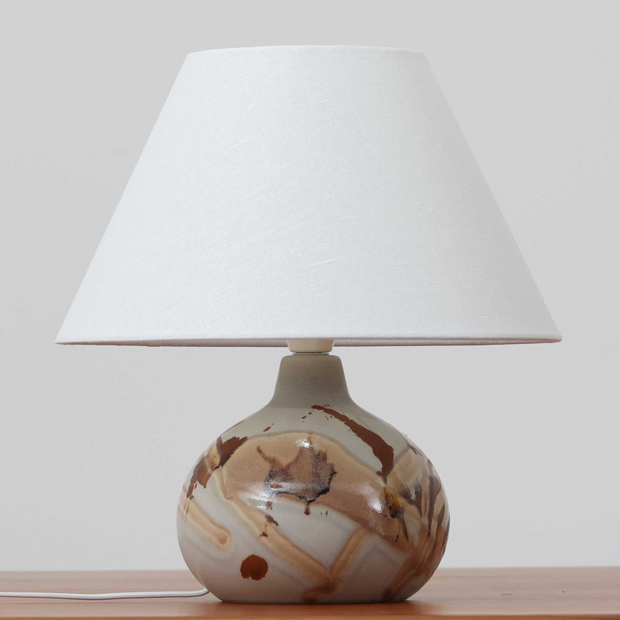 Soholm Keramik（スーホルム・セラミック）が手掛けたこちらのテーブルランプは、淡いグレーを基調にブラウンの流れるような釉薬模様が美しく広がる有機的な表情が魅力で、丸みを帯びた陶磁器のベースが空間にやさしい存在感をもたらす北欧ビンテージ品です。リネンシェードを透ける柔らかな灯りが落ち着いた雰囲気をつくり、ベッドサイドやリビングの間接照明として取り入れやすく、経年によって深まった色合いと一点物ならではの味わいを長く楽しめる一台です。