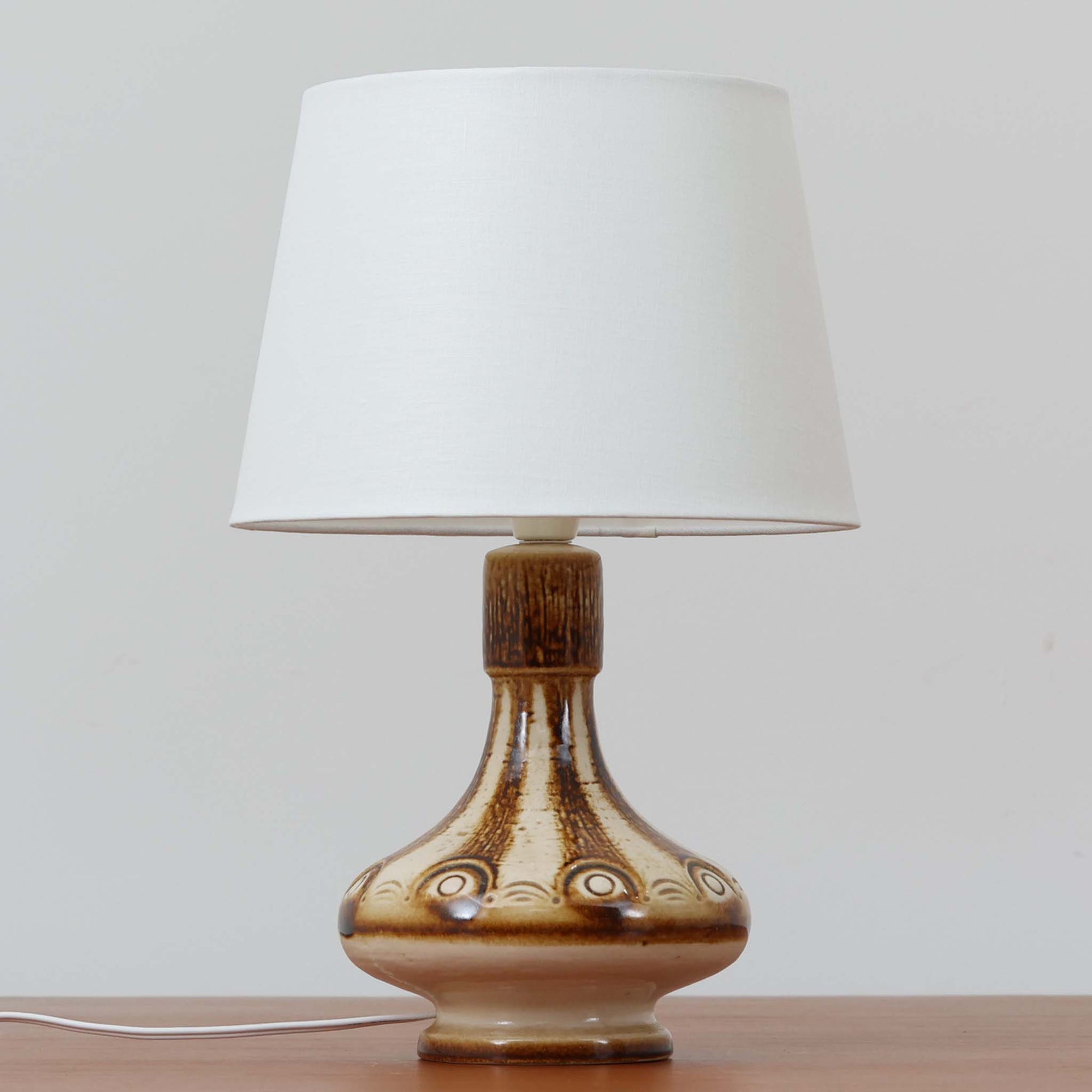 Soholm Keramik（スーホルム・セラミック）が手掛けたこちらのテーブルランプは、柔らかなクリーム色を基調にブラウンの縦流れ模様と円形のレリーフ装飾があしらわれた温かみある陶磁器ベースが魅力で、ふっくらと広がるフォルムが北欧ビンテージらしいやさしい存在感を放つ一台です。リネンシェード越しに灯る穏やかな光はベッドサイドやリビングの間接照明として心地よく空間を包み込み、経年で深まった釉薬の表情が一点物ならではの味わいを引き立てています。