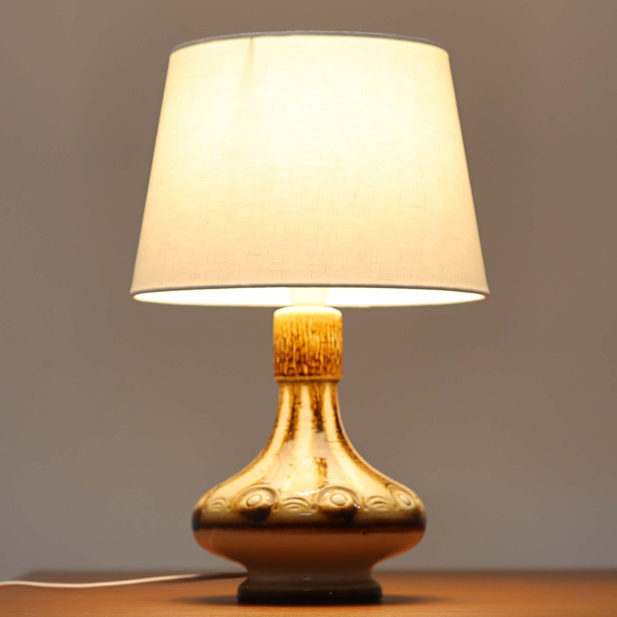 Soholm Keramik（スーホルム・セラミック）が手掛けたこちらのテーブルランプは、柔らかなクリーム色を基調にブラウンの縦流れ模様と円形のレリーフ装飾があしらわれた温かみある陶磁器ベースが魅力で、ふっくらと広がるフォルムが北欧ビンテージらしいやさしい存在感を放つ一台です。リネンシェード越しに灯る穏やかな光はベッドサイドやリビングの間接照明として心地よく空間を包み込み、経年で深まった釉薬の表情が一点物ならではの味わいを引き立てています。