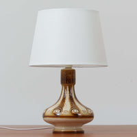 Soholm Keramik（スーホルム・セラミック）が手掛けたこちらのテーブルランプは、柔らかなクリーム色を基調にブラウンの縦流れ模様と円形のレリーフ装飾があしらわれた温かみある陶磁器ベースが魅力で、ふっくらと広がるフォルムが北欧ビンテージらしいやさしい存在感を放つ一台です。リネンシェード越しに灯る穏やかな光はベッドサイドやリビングの間接照明として心地よく空間を包み込み、経年で深まった釉薬の表情が一点物ならではの味わいを引き立てています。