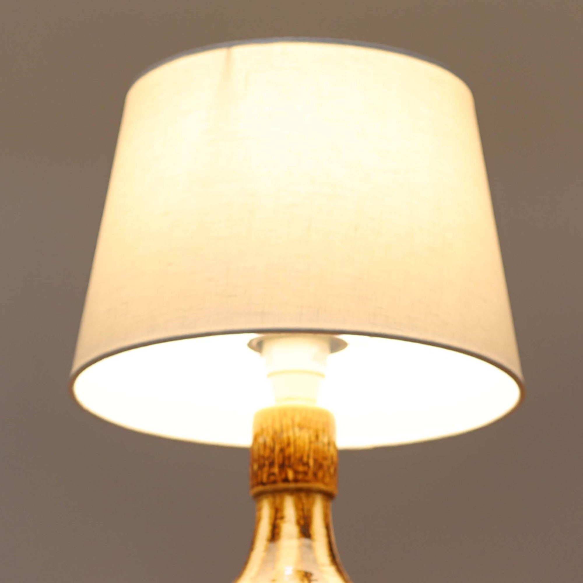 Soholm Keramik（スーホルム・セラミック）が手掛けたこちらのテーブルランプは、柔らかなクリーム色を基調にブラウンの縦流れ模様と円形のレリーフ装飾があしらわれた温かみある陶磁器ベースが魅力で、ふっくらと広がるフォルムが北欧ビンテージらしいやさしい存在感を放つ一台です。リネンシェード越しに灯る穏やかな光はベッドサイドやリビングの間接照明として心地よく空間を包み込み、経年で深まった釉薬の表情が一点物ならではの味わいを引き立てています。