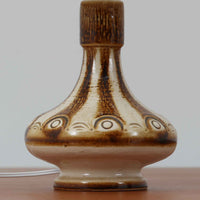 Soholm Keramik（スーホルム・セラミック）が手掛けたこちらのテーブルランプは、柔らかなクリーム色を基調にブラウンの縦流れ模様と円形のレリーフ装飾があしらわれた温かみある陶磁器ベースが魅力で、ふっくらと広がるフォルムが北欧ビンテージらしいやさしい存在感を放つ一台です。リネンシェード越しに灯る穏やかな光はベッドサイドやリビングの間接照明として心地よく空間を包み込み、経年で深まった釉薬の表情が一点物ならではの味わいを引き立てています。