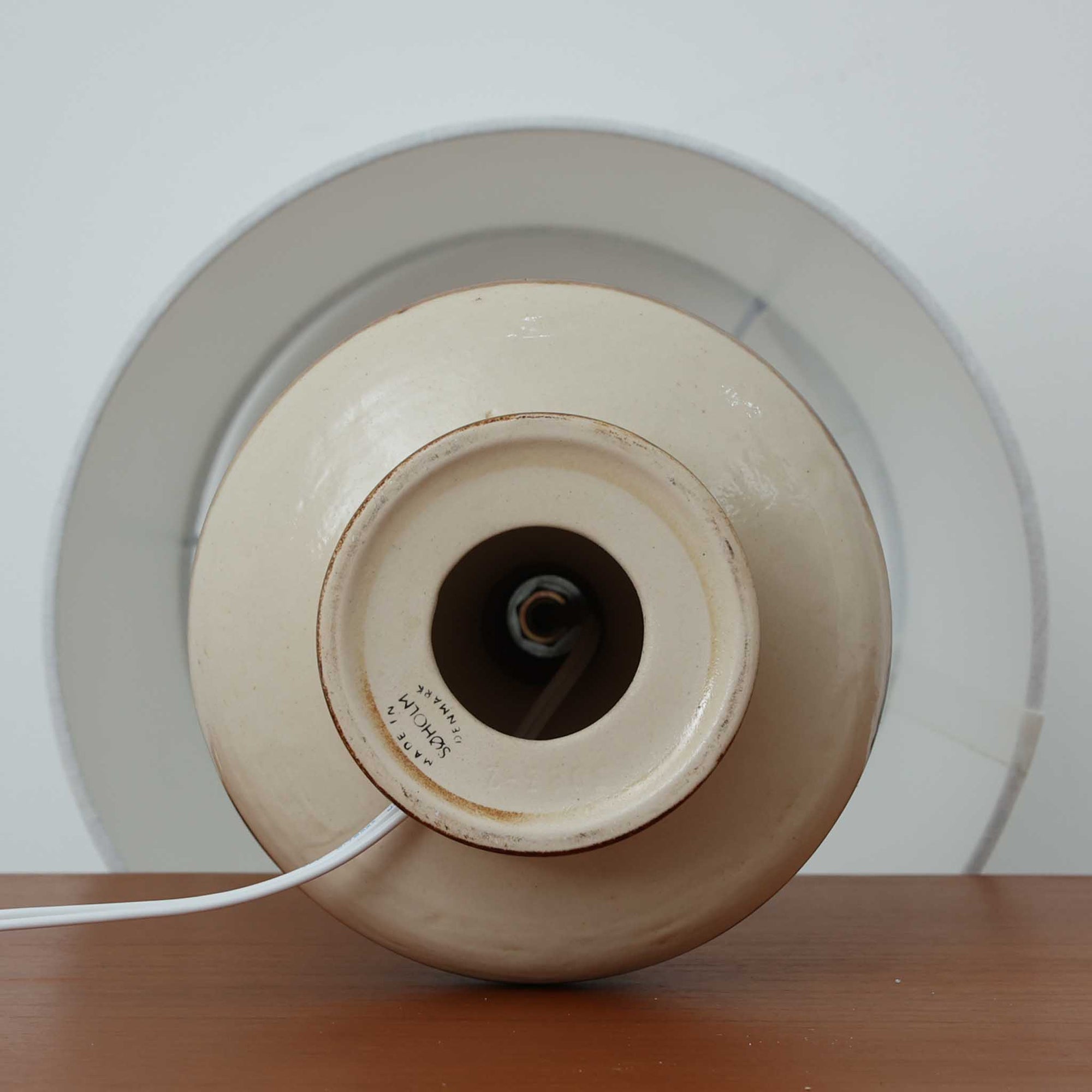 Soholm Keramik（スーホルム・セラミック）が手掛けたこちらのテーブルランプは、柔らかなクリーム色を基調にブラウンの縦流れ模様と円形のレリーフ装飾があしらわれた温かみある陶磁器ベースが魅力で、ふっくらと広がるフォルムが北欧ビンテージらしいやさしい存在感を放つ一台です。リネンシェード越しに灯る穏やかな光はベッドサイドやリビングの間接照明として心地よく空間を包み込み、経年で深まった釉薬の表情が一点物ならではの味わいを引き立てています。