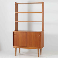 スウェーデン製のこちらのオープンシェルフは、チーク材の温かな木目とオーク材の脚部を組み合わせた軽やかな佇まいが魅力で、上段の棚板と下段のスライド扉収納を備えた実用性の高い構成が特徴の北欧ビンテージ家具です。丸い掘り込み取手や細身のフレームなど繊細なディテールが空間を柔らかく彩り、本や雑貨、食器などを美しく見せながら整頓でき、経年で深まった木肌の風合いがインテリアに落ち着きと温もりを与える一台です。