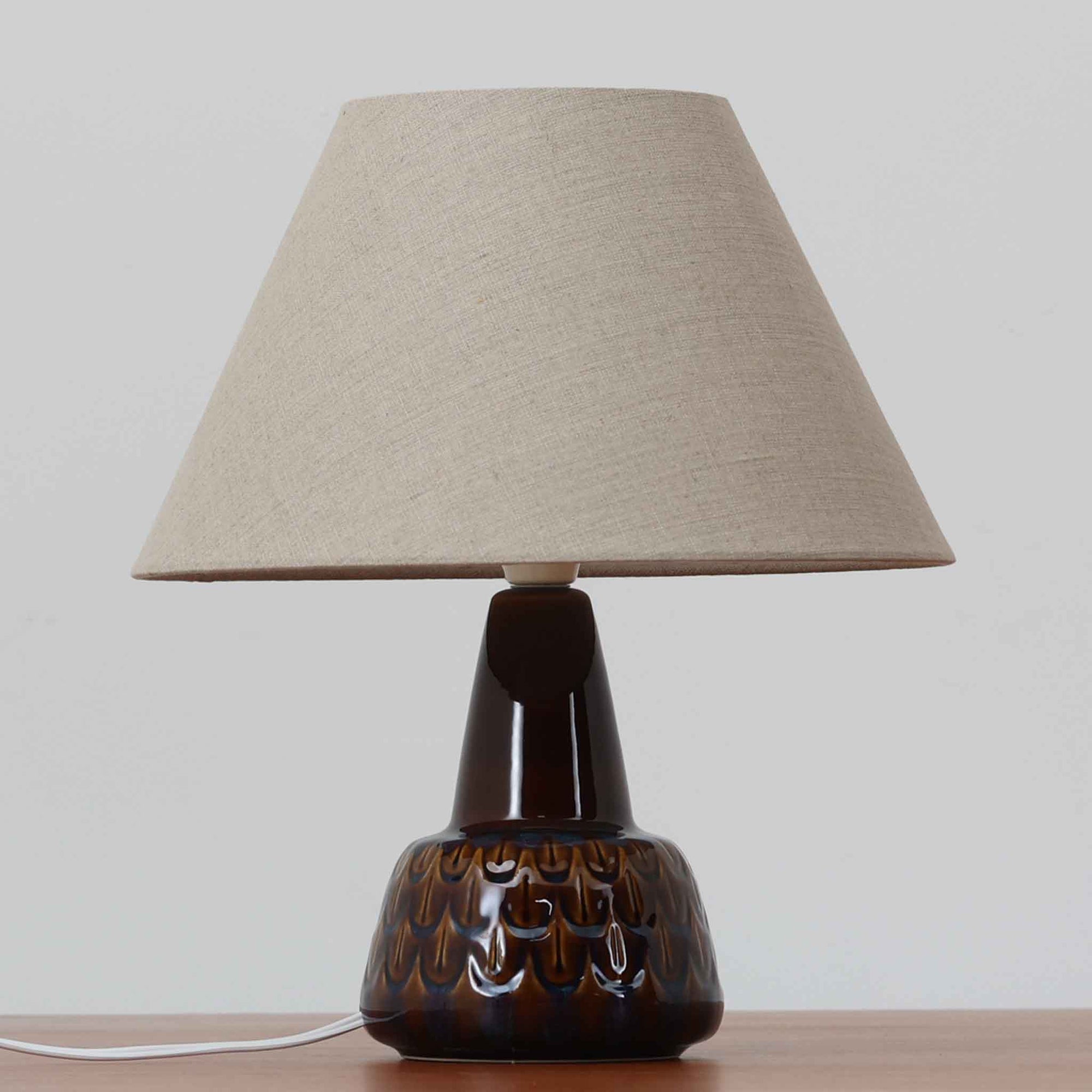 Einar JohansenがSoholm Keramik（スーホルム・セラミック）のために手掛けたモデル1015は、深いブラウンの釉薬に美しい光沢が宿り、ベースを覆う羽根模様のようなレリーフが立体的な表情を生み出す北欧ビンテージの名品で、すっきりと立ち上がるシルエットと陶磁器ならではの重厚感が空間に落ち着いた存在感をもたらします。リネンシェードを通したやわらかな灯りはベッドサイドやリビングに 温かみを添え、経年の風合いが一点物としての魅力をより一層引き立てています。