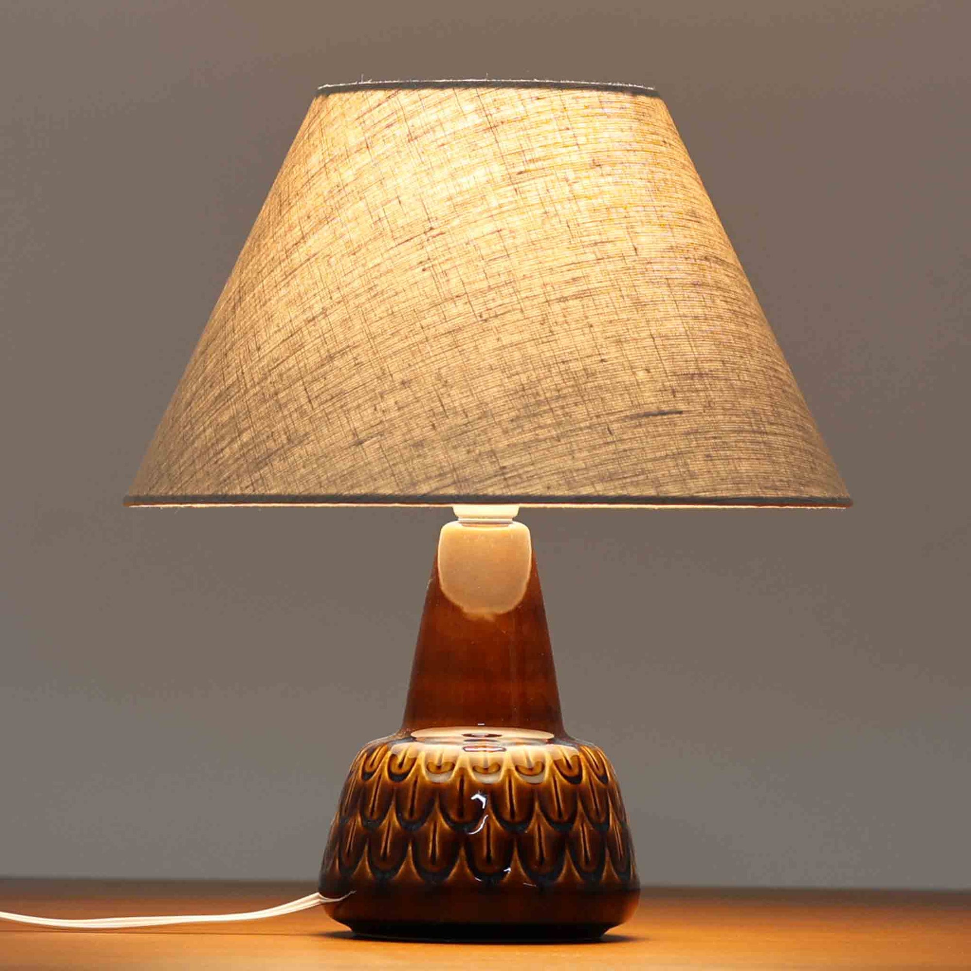 Einar JohansenがSoholm Keramik（スーホルム・セラミック）のために手掛けたモデル1015は、深いブラウンの釉薬に美しい光沢が宿り、ベースを覆う羽根模様のようなレリーフが立体的な表情を生み出す北欧ビンテージの名品で、すっきりと立ち上がるシルエットと陶磁器ならではの重厚感が空間に落ち着いた存在感をもたらします。リネンシェードを通したやわらかな灯りはベッドサイドやリビングに 温かみを添え、経年の風合いが一点物としての魅力をより一層引き立てています。