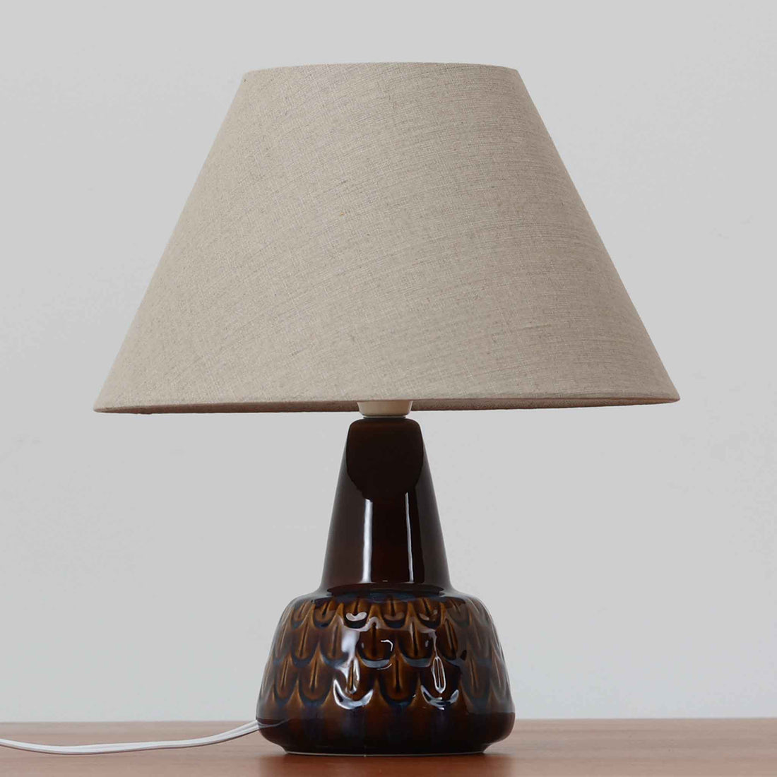 Einar JohansenがSoholm Keramik（スーホルム・セラミック）のために手掛けたモデル1015は、深いブラウンの釉薬に美しい光沢が宿り、ベースを覆う羽根模様のようなレリーフが立体的な表情を生み出す北欧ビンテージの名品で、すっきりと立ち上がるシルエットと陶磁器ならではの重厚感が空間に落ち着いた存在感をもたらします。リネンシェードを通したやわらかな灯りはベッドサイドやリビングに 温かみを添え、経年の風合いが一点物としての魅力をより一層引き立てています。