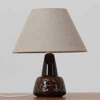 Einar JohansenがSoholm Keramik（スーホルム・セラミック）のために手掛けたモデル1015は、深いブラウンの釉薬に美しい光沢が宿り、ベースを覆う羽根模様のようなレリーフが立体的な表情を生み出す北欧ビンテージの名品で、すっきりと立ち上がるシルエットと陶磁器ならではの重厚感が空間に落ち着いた存在感をもたらします。リネンシェードを通したやわらかな灯りはベッドサイドやリビングに 温かみを添え、経年の風合いが一点物としての魅力をより一層引き立てています。