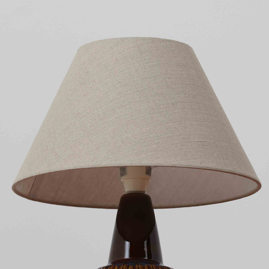 Einar JohansenがSoholm Keramik（スーホルム・セラミック）のために手掛けたモデル1015は、深いブラウンの釉薬に美しい光沢が宿り、ベースを覆う羽根模様のようなレリーフが立体的な表情を生み出す北欧ビンテージの名品で、すっきりと立ち上がるシルエットと陶磁器ならではの重厚感が空間に落ち着いた存在感をもたらします。リネンシェードを通したやわらかな灯りはベッドサイドやリビングに 温かみを添え、経年の風合いが一点物としての魅力をより一層引き立てています。