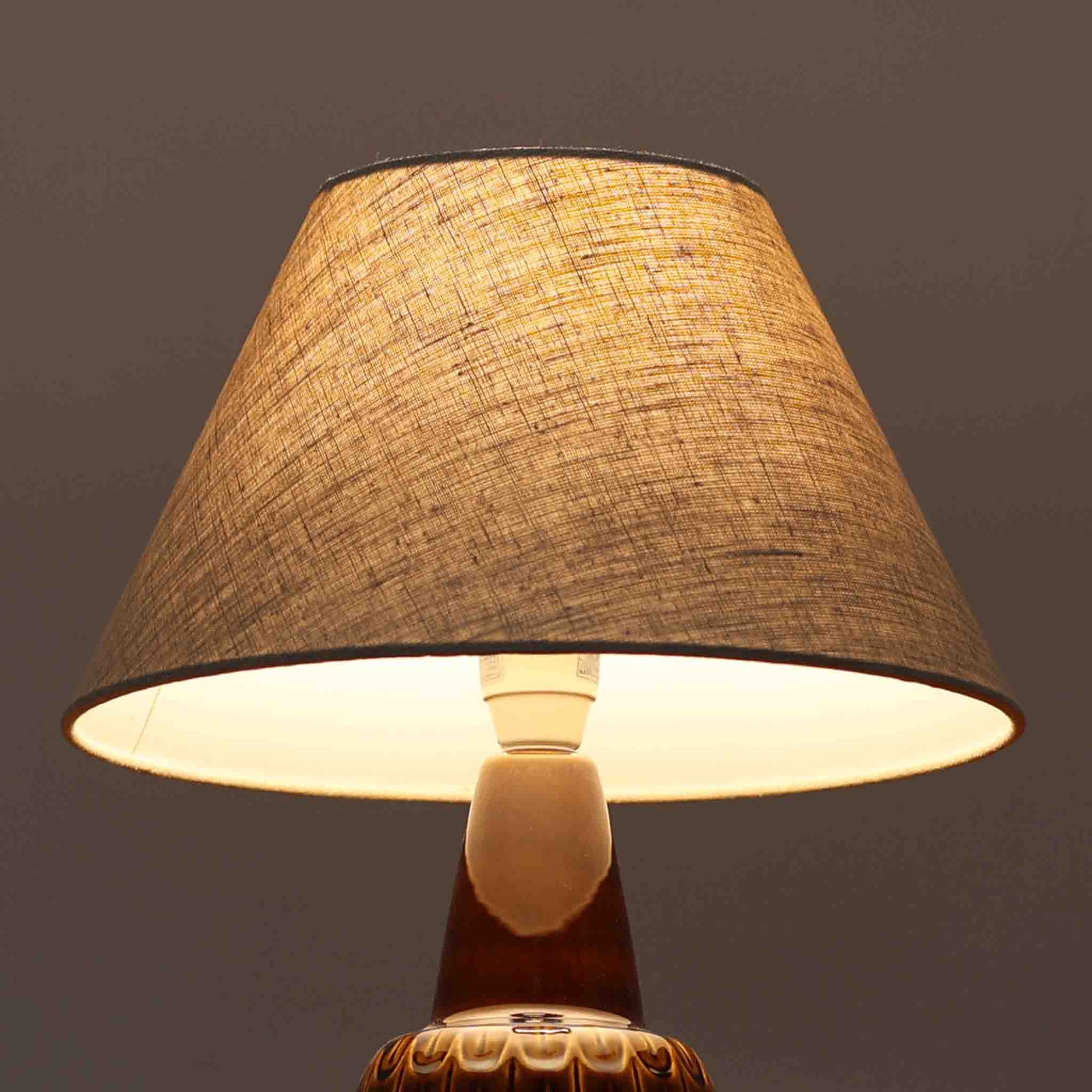 Einar JohansenがSoholm Keramik（スーホルム・セラミック）のために手掛けたモデル1015は、深いブラウンの釉薬に美しい光沢が宿り、ベースを覆う羽根模様のようなレリーフが立体的な表情を生み出す北欧ビンテージの名品で、すっきりと立ち上がるシルエットと陶磁器ならではの重厚感が空間に落ち着いた存在感をもたらします。リネンシェードを通したやわらかな灯りはベッドサイドやリビングに 温かみを添え、経年の風合いが一点物としての魅力をより一層引き立てています。