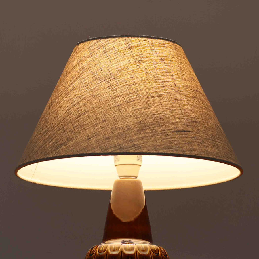 Einar JohansenがSoholm Keramik（スーホルム・セラミック）のために手掛けたモデル1015は、深いブラウンの釉薬に美しい光沢が宿り、ベースを覆う羽根模様のようなレリーフが立体的な表情を生み出す北欧ビンテージの名品で、すっきりと立ち上がるシルエットと陶磁器ならではの重厚感が空間に落ち着いた存在感をもたらします。リネンシェードを通したやわらかな灯りはベッドサイドやリビングに 温かみを添え、経年の風合いが一点物としての魅力をより一層引き立てています。