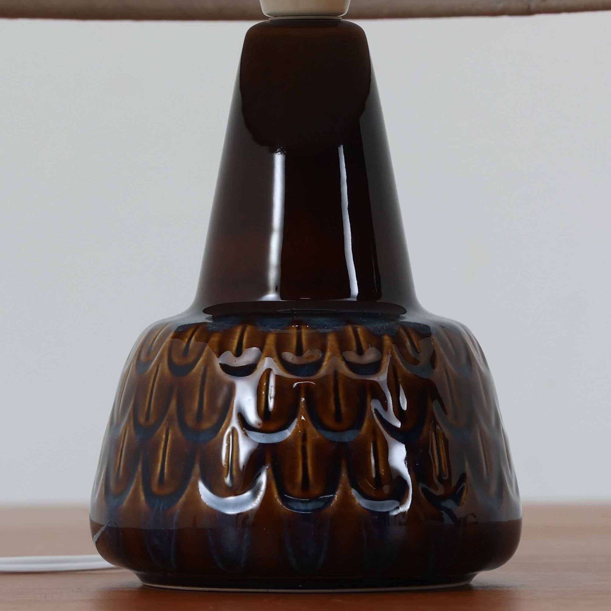 Einar JohansenがSoholm Keramik（スーホルム・セラミック）のために手掛けたモデル1015は、深いブラウンの釉薬に美しい光沢が宿り、ベースを覆う羽根模様のようなレリーフが立体的な表情を生み出す北欧ビンテージの名品で、すっきりと立ち上がるシルエットと陶磁器ならではの重厚感が空間に落ち着いた存在感をもたらします。リネンシェードを通したやわらかな灯りはベッドサイドやリビングに 温かみを添え、経年の風合いが一点物としての魅力をより一層引き立てています。