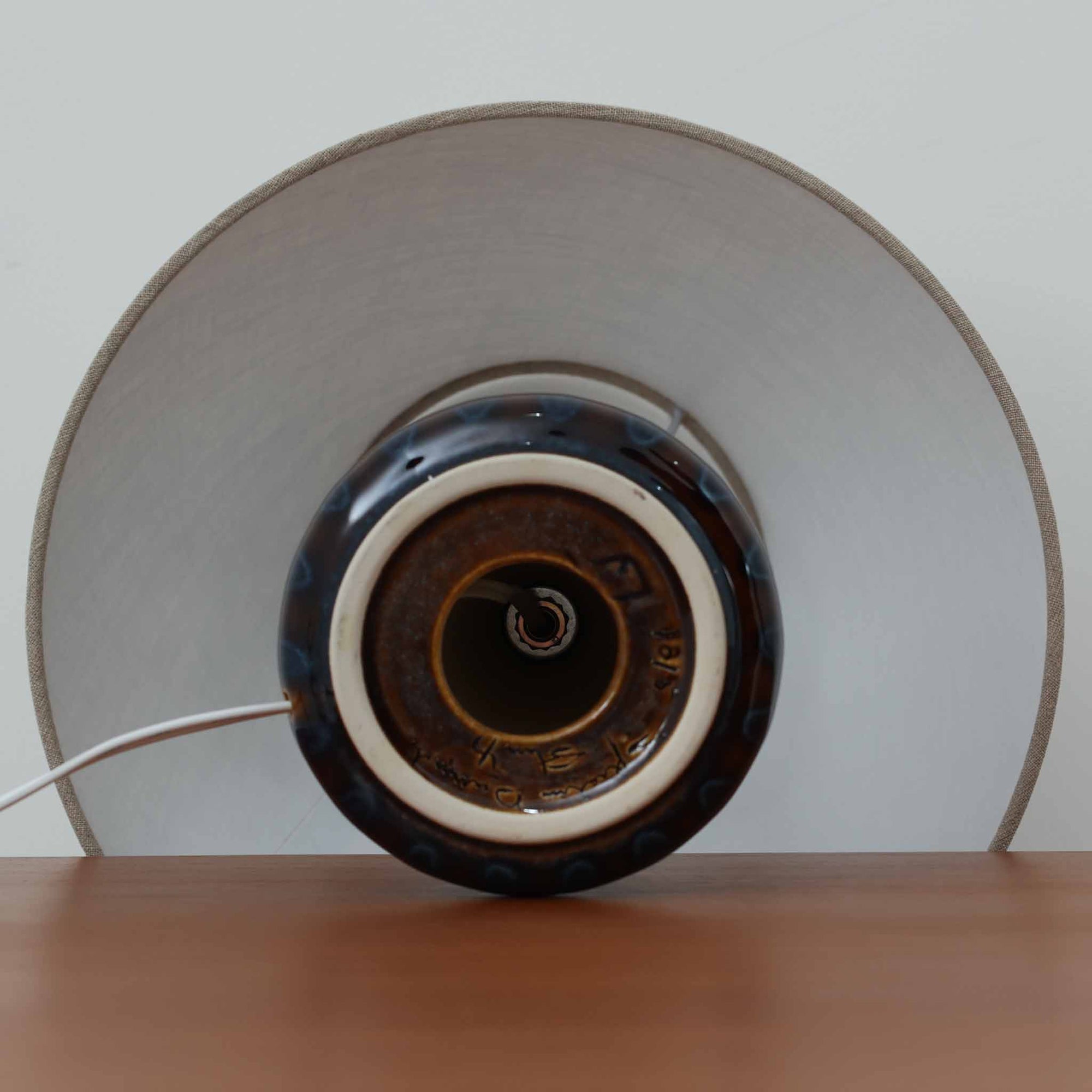 Einar JohansenがSoholm Keramik（スーホルム・セラミック）のために手掛けたモデル1015は、深いブラウンの釉薬に美しい光沢が宿り、ベースを覆う羽根模様のようなレリーフが立体的な表情を生み出す北欧ビンテージの名品で、すっきりと立ち上がるシルエットと陶磁器ならではの重厚感が空間に落ち着いた存在感をもたらします。リネンシェードを通したやわらかな灯りはベッドサイドやリビングに 温かみを添え、経年の風合いが一点物としての魅力をより一層引き立てています。