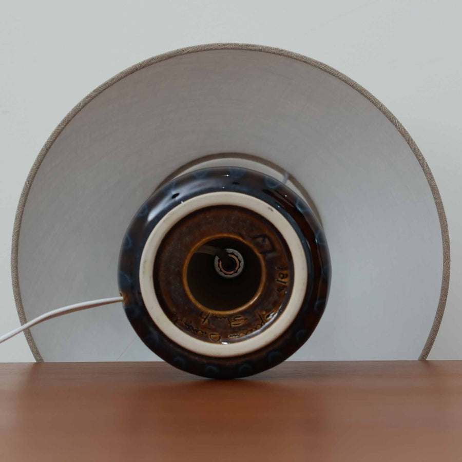 Einar JohansenがSoholm Keramik（スーホルム・セラミック）のために手掛けたモデル1015は、深いブラウンの釉薬に美しい光沢が宿り、ベースを覆う羽根模様のようなレリーフが立体的な表情を生み出す北欧ビンテージの名品で、すっきりと立ち上がるシルエットと陶磁器ならではの重厚感が空間に落ち着いた存在感をもたらします。リネンシェードを通したやわらかな灯りはベッドサイドやリビングに 温かみを添え、経年の風合いが一点物としての魅力をより一層引き立てています。