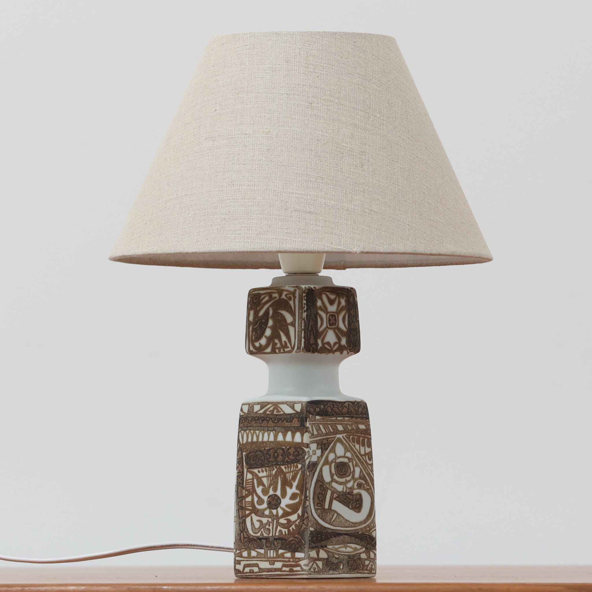 Nils Thorsson（ニルス・トールソン）がRoyal Copenhagen（ロイヤルコペンハーゲン）のために手掛けた「BACA」シリーズのテーブルランプは、繊細な線描と象徴的なモチーフが幾何学的に配置された陶磁器ベースが特徴で、手仕事ならではの奥行きあるブラウンの釉調がビンテージとしての存在感を高めています。上下で形状が切り替わる立体的な構造が光を受けて表情を変え、リネンシェードを通した柔らかな灯りが空間に落ち着いた陰影を添えます。高さ36cmの扱いやすいサイズで、リビングや寝室のサイドテーブルにもよく馴染み、北欧デザインの芸術性と実用性を兼ね備えた一台です。