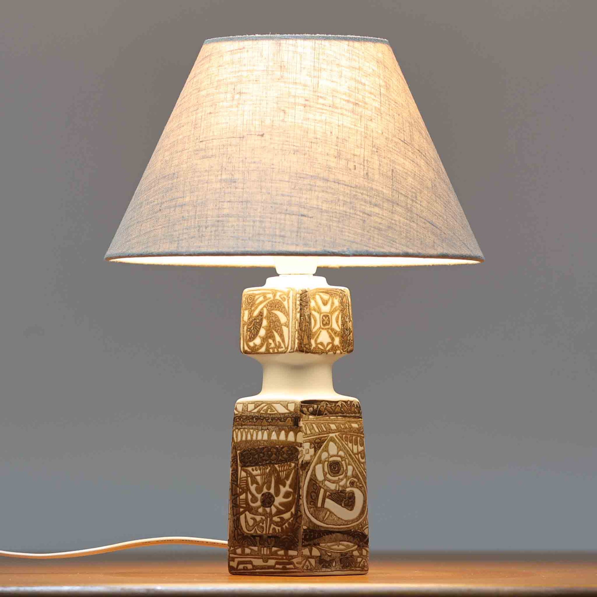 Nils Thorsson（ニルス・トールソン）がRoyal Copenhagen（ロイヤルコペンハーゲン）のために手掛けた「BACA」シリーズのテーブルランプは、繊細な線描と象徴的なモチーフが幾何学的に配置された陶磁器ベースが特徴で、手仕事ならではの奥行きあるブラウンの釉調がビンテージとしての存在感を高めています。上下で形状が切り替わる立体的な構造が光を受けて表情を変え、リネンシェードを通した柔らかな灯りが空間に落ち着いた陰影を添えます。高さ36cmの扱いやすいサイズで、リビングや寝室のサイドテーブルにもよく馴染み、北欧デザインの芸術性と実用性を兼ね備えた一台です。