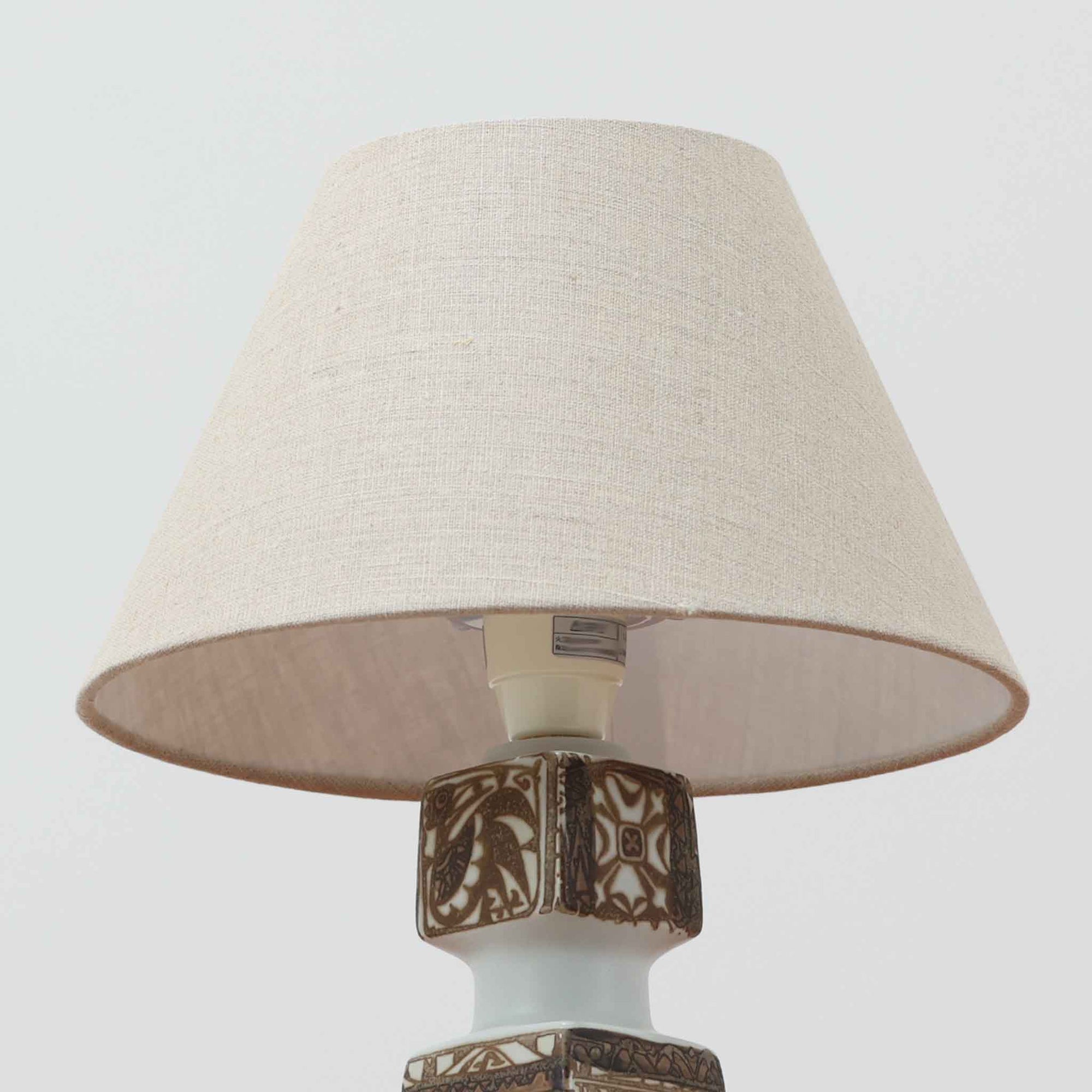 Nils Thorsson（ニルス・トールソン）がRoyal Copenhagen（ロイヤルコペンハーゲン）のために手掛けた「BACA」シリーズのテーブルランプは、繊細な線描と象徴的なモチーフが幾何学的に配置された陶磁器ベースが特徴で、手仕事ならではの奥行きあるブラウンの釉調がビンテージとしての存在感を高めています。上下で形状が切り替わる立体的な構造が光を受けて表情を変え、リネンシェードを通した柔らかな灯りが空間に落ち着いた陰影を添えます。高さ36cmの扱いやすいサイズで、リビングや寝室のサイドテーブルにもよく馴染み、北欧デザインの芸術性と実用性を兼ね備えた一台です。