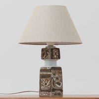 Nils Thorsson（ニルス・トールソン）がRoyal Copenhagen（ロイヤルコペンハーゲン）のために手掛けた「BACA」シリーズのテーブルランプは、繊細な線描と象徴的なモチーフが幾何学的に配置された陶磁器ベースが特徴で、手仕事ならではの奥行きあるブラウンの釉調がビンテージとしての存在感を高めています。上下で形状が切り替わる立体的な構造が光を受けて表情を変え、リネンシェードを通した柔らかな灯りが空間に落ち着いた陰影を添えます。高さ36cmの扱いやすいサイズで、リビングや寝室のサイドテーブルにもよく馴染み、北欧デザインの芸術性と実用性を兼ね備えた一台です。