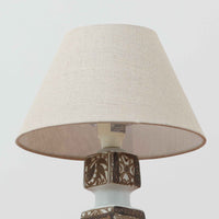 Nils Thorsson（ニルス・トールソン）がRoyal Copenhagen（ロイヤルコペンハーゲン）のために手掛けた「BACA」シリーズのテーブルランプは、繊細な線描と象徴的なモチーフが幾何学的に配置された陶磁器ベースが特徴で、手仕事ならではの奥行きあるブラウンの釉調がビンテージとしての存在感を高めています。上下で形状が切り替わる立体的な構造が光を受けて表情を変え、リネンシェードを通した柔らかな灯りが空間に落ち着いた陰影を添えます。高さ36cmの扱いやすいサイズで、リビングや寝室のサイドテーブルにもよく馴染み、北欧デザインの芸術性と実用性を兼ね備えた一台です。