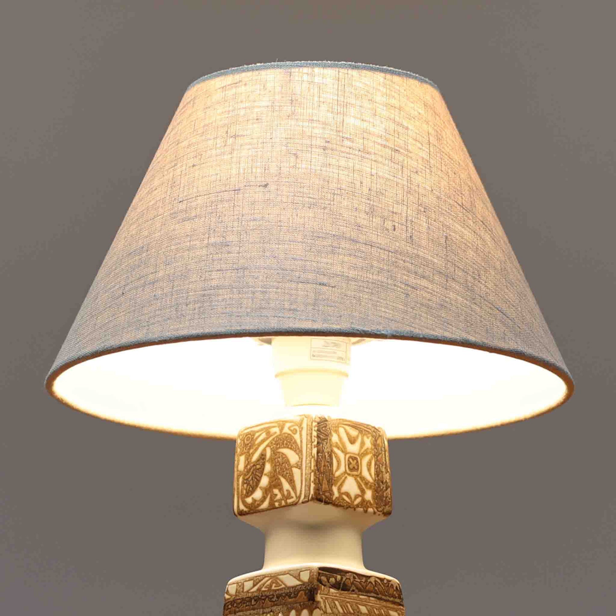 Nils Thorsson（ニルス・トールソン）がRoyal Copenhagen（ロイヤルコペンハーゲン）のために手掛けた「BACA」シリーズのテーブルランプは、繊細な線描と象徴的なモチーフが幾何学的に配置された陶磁器ベースが特徴で、手仕事ならではの奥行きあるブラウンの釉調がビンテージとしての存在感を高めています。上下で形状が切り替わる立体的な構造が光を受けて表情を変え、リネンシェードを通した柔らかな灯りが空間に落ち着いた陰影を添えます。高さ36cmの扱いやすいサイズで、リビングや寝室のサイドテーブルにもよく馴染み、北欧デザインの芸術性と実用性を兼ね備えた一台です。