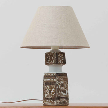 Nils Thorsson（ニルス・トールソン）がRoyal Copenhagen（ロイヤルコペンハーゲン）のために手掛けた「BACA」シリーズのテーブルランプは、繊細な線描と象徴的なモチーフが幾何学的に配置された陶磁器ベースが特徴で、手仕事ならではの奥行きあるブラウンの釉調がビンテージとしての存在感を高めています。上下で形状が切り替わる立体的な構造が光を受けて表情を変え、リネンシェードを通した柔らかな灯りが空間に落ち着いた陰影を添えます。高さ36cmの扱いやすいサイズで、リビングや寝室のサイドテーブルにもよく馴染み、北欧デザインの芸術性と実用性を兼ね備えた一台です。