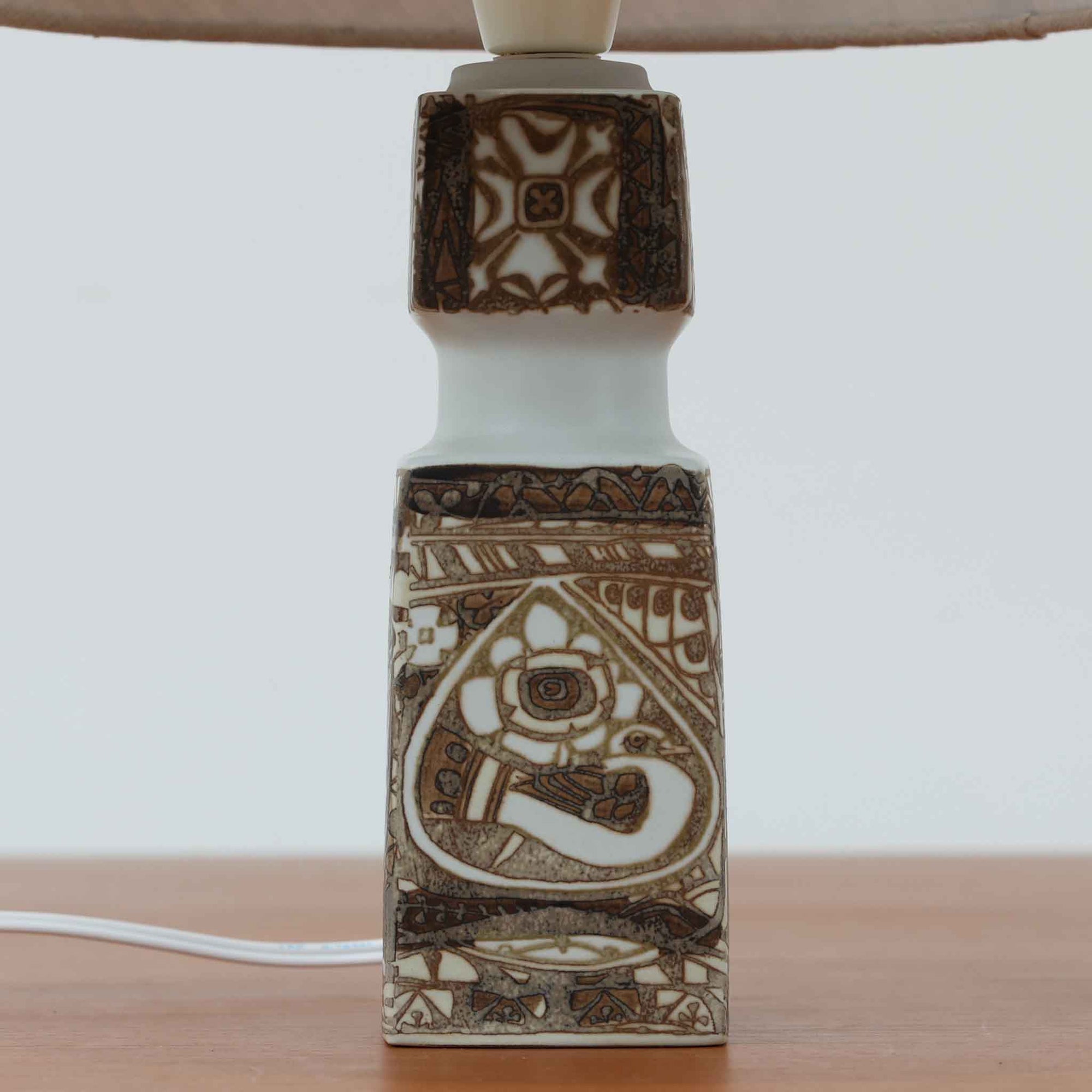 Nils Thorsson（ニルス・トールソン）がRoyal Copenhagen（ロイヤルコペンハーゲン）のために手掛けた「BACA」シリーズのテーブルランプは、繊細な線描と象徴的なモチーフが幾何学的に配置された陶磁器ベースが特徴で、手仕事ならではの奥行きあるブラウンの釉調がビンテージとしての存在感を高めています。上下で形状が切り替わる立体的な構造が光を受けて表情を変え、リネンシェードを通した柔らかな灯りが空間に落ち着いた陰影を添えます。高さ36cmの扱いやすいサイズで、リビングや寝室のサイドテーブルにもよく馴染み、北欧デザインの芸術性と実用性を兼ね備えた一台です。