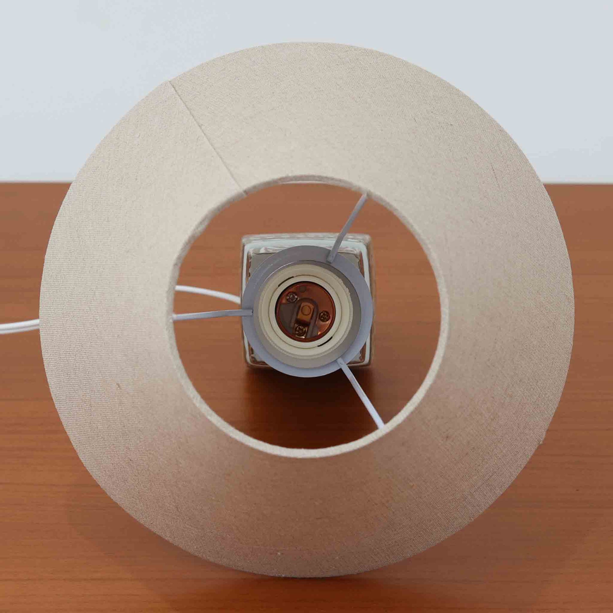 Nils Thorsson（ニルス・トールソン）がRoyal Copenhagen（ロイヤルコペンハーゲン）のために手掛けた「BACA」シリーズのテーブルランプは、繊細な線描と象徴的なモチーフが幾何学的に配置された陶磁器ベースが特徴で、手仕事ならではの奥行きあるブラウンの釉調がビンテージとしての存在感を高めています。上下で形状が切り替わる立体的な構造が光を受けて表情を変え、リネンシェードを通した柔らかな灯りが空間に落ち着いた陰影を添えます。高さ36cmの扱いやすいサイズで、リビングや寝室のサイドテーブルにもよく馴染み、北欧デザインの芸術性と実用性を兼ね備えた一台です。