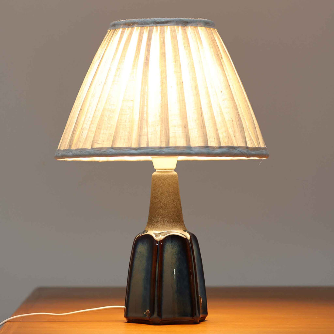 Einar JohansenがSoholm Keramik（スーホルム・セラミック）のためにデザインしたモデル1031は、深みのあるブルーの釉薬と立体的な面取りが織りなす陰影が美しい陶磁器ベースが特徴で、上部のマットな質感との対比がモダンで彫刻的な存在感を放つ北欧ビンテージ品です。プリーツシェードを透ける柔らかな灯りが落ち着いた雰囲気をつくり、ベッドサイドやリビングのアクセント照明として取り入れやすく、経年で深まった釉薬の表情が一点物ならではの魅力を一層引き立てています。