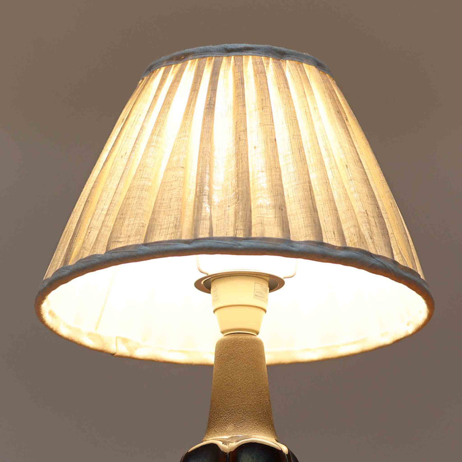 Einar JohansenがSoholm Keramik（スーホルム・セラミック）のためにデザインしたモデル1031は、深みのあるブルーの釉薬と立体的な面取りが織りなす陰影が美しい陶磁器ベースが特徴で、上部のマットな質感との対比がモダンで彫刻的な存在感を放つ北欧ビンテージ品です。プリーツシェードを透ける柔らかな灯りが落ち着いた雰囲気をつくり、ベッドサイドやリビングのアクセント照明として取り入れやすく、経年で深まった釉薬の表情が一点物ならではの魅力を一層引き立てています。