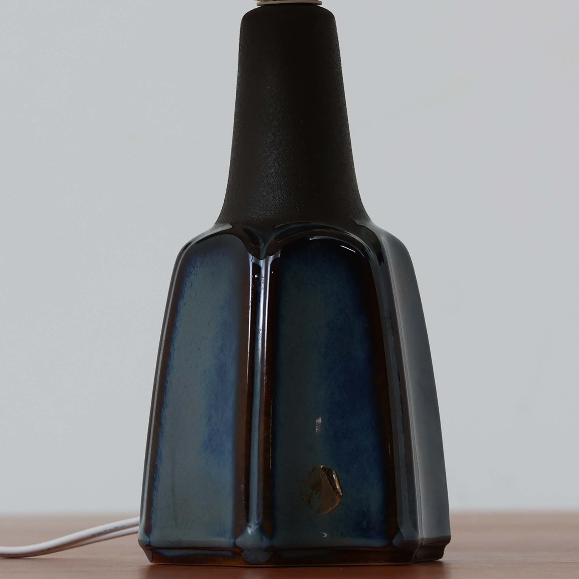 Einar JohansenがSoholm Keramik（スーホルム・セラミック）のためにデザインしたモデル1031は、深みのあるブルーの釉薬と立体的な面取りが織りなす陰影が美しい陶磁器ベースが特徴で、上部のマットな質感との対比がモダンで彫刻的な存在感を放つ北欧ビンテージ品です。プリーツシェードを透ける柔らかな灯りが落ち着いた雰囲気をつくり、ベッドサイドやリビングのアクセント照明として取り入れやすく、経年で深まった釉薬の表情が一点物ならではの魅力を一層引き立てています。