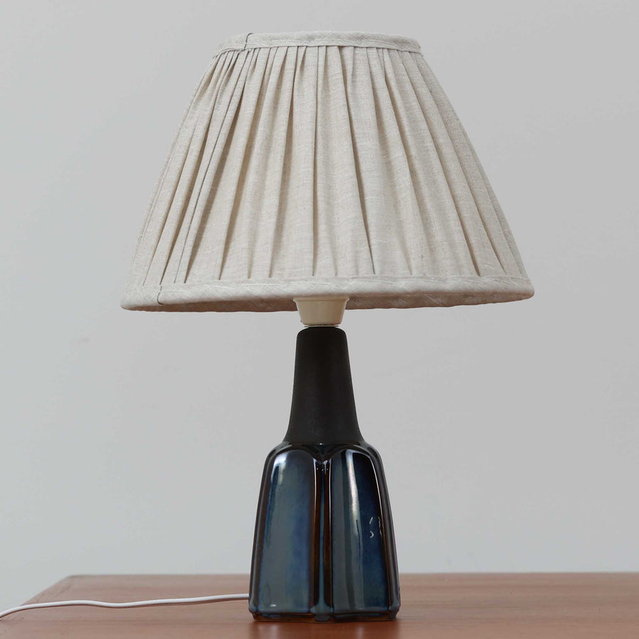 Einar JohansenがSoholm Keramik（スーホルム・セラミック）のためにデザインしたモデル1031は、深みのあるブルーの釉薬と立体的な面取りが織りなす陰影が美しい陶磁器ベースが特徴で、上部のマットな質感との対比がモダンで彫刻的な存在感を放つ北欧ビンテージ品です。プリーツシェードを透ける柔らかな灯りが落ち着いた雰囲気をつくり、ベッドサイドやリビングのアクセント照明として取り入れやすく、経年で深まった釉薬の表情が一点物ならではの魅力を一層引き立てています。