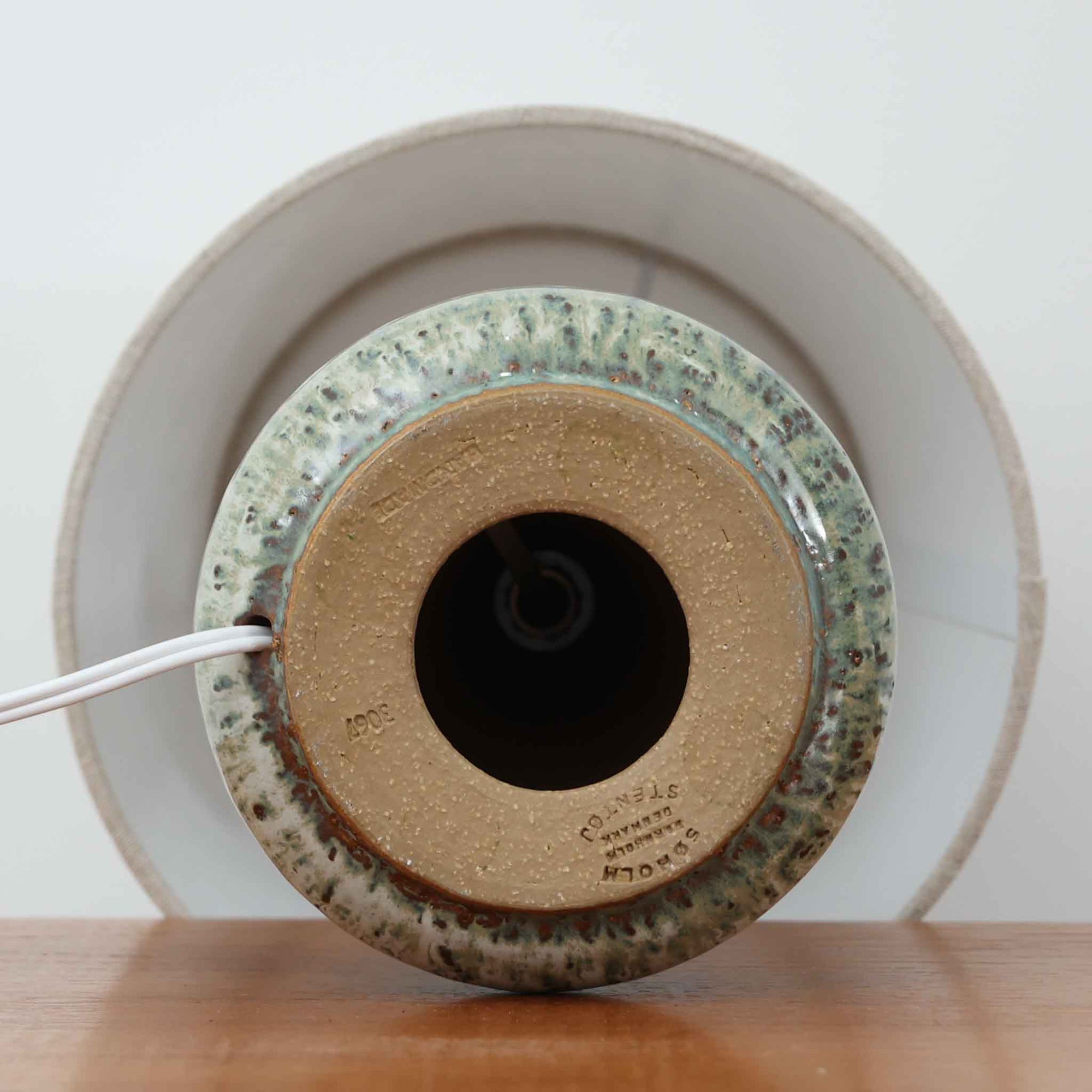 Soholm Keramikが手掛けたモデル3067のテーブルランプです。丸みを帯びた安定感のある陶磁器ベースから、すっと立ち上がる首元のフォルムが美しく、深みのあるグリーンを基調にした流れるような釉薬の景色が一点物のような存在感を放ちます。リネンシェードを通した柔らかな光が陶器の艶やかな質感を引き立て、サイドテーブルやチェスト上、ベッドサイドに落ち着いた北欧らしい温もりを添えてくれる一台。経年で味わいを増した釉薬の表情と職人の手仕事を感じる焼きの風合いが、ビンテージならではの希少な魅力を高めています。