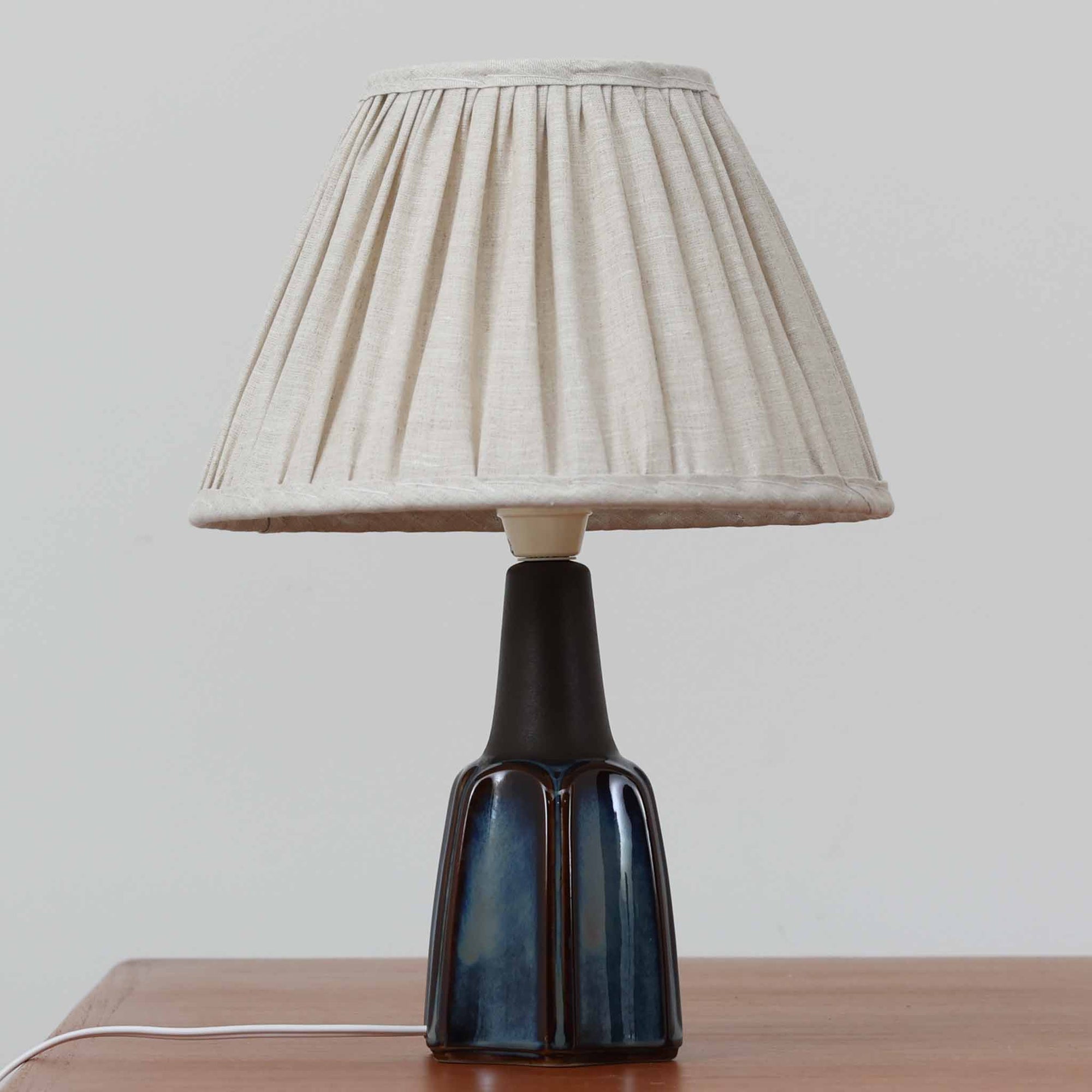 Einar JohansenがSoholm Keramik（スーホルム・セラミック）のためにデザインしたモデル1031は、深みのあるブルーの釉薬と立体的な面取りが織りなす陰影が美しい陶磁器ベースが特徴で、上部のマットな質感との対比がモダンで彫刻的な存在感を放つ北欧ビンテージ品です。プリーツシェードを透ける柔らかな灯りが落ち着いた雰囲気をつくり、ベッドサイドやリビングのアクセント照明として取り入れやすく、経年で深まった釉薬の表情が一点物ならではの魅力を一層引き立てています。