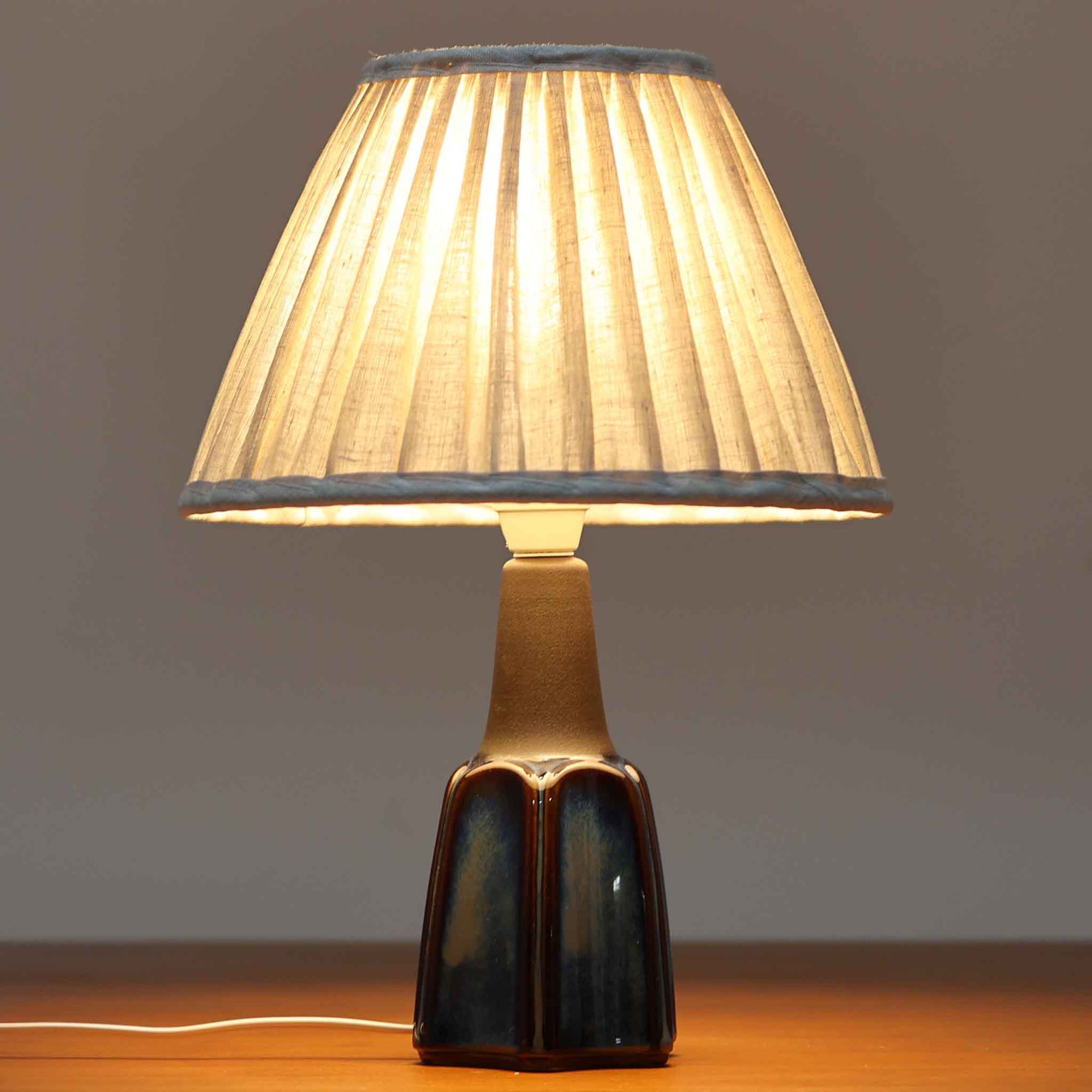 Einar JohansenがSoholm Keramik（スーホルム・セラミック）のためにデザインしたモデル1031は、深みのあるブルーの釉薬と立体的な面取りが織りなす陰影が美しい陶磁器ベースが特徴で、上部のマットな質感との対比がモダンで彫刻的な存在感を放つ北欧ビンテージ品です。プリーツシェードを透ける柔らかな灯りが落ち着いた雰囲気をつくり、ベッドサイドやリビングのアクセント照明として取り入れやすく、経年で深まった釉薬の表情が一点物ならではの魅力を一層引き立てています。