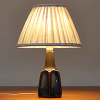 Einar JohansenがSoholm Keramik（スーホルム・セラミック）のためにデザインしたモデル1031は、深みのあるブルーの釉薬と立体的な面取りが織りなす陰影が美しい陶磁器ベースが特徴で、上部のマットな質感との対比がモダンで彫刻的な存在感を放つ北欧ビンテージ品です。プリーツシェードを透ける柔らかな灯りが落ち着いた雰囲気をつくり、ベッドサイドやリビングのアクセント照明として取り入れやすく、経年で深まった釉薬の表情が一点物ならではの魅力を一層引き立てています。