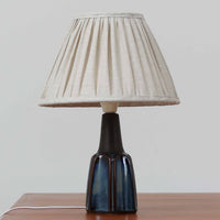 Einar JohansenがSoholm Keramik（スーホルム・セラミック）のためにデザインしたモデル1031は、深みのあるブルーの釉薬と立体的な面取りが織りなす陰影が美しい陶磁器ベースが特徴で、上部のマットな質感との対比がモダンで彫刻的な存在感を放つ北欧ビンテージ品です。プリーツシェードを透ける柔らかな灯りが落ち着いた雰囲気をつくり、ベッドサイドやリビングのアクセント照明として取り入れやすく、経年で深まった釉薬の表情が一点物ならではの魅力を一層引き立てています。